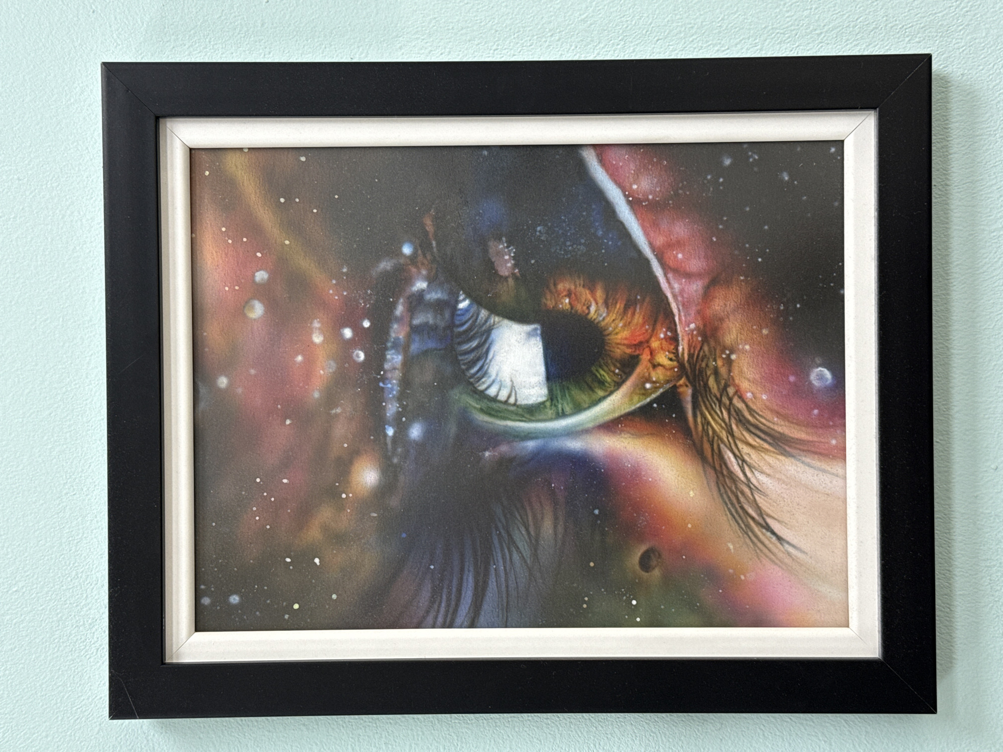 A framed painting of an eye, with a celestial background, hangs on a pale blue wall. The frame is black with a white inner border. The painting dominates the field of view, showing a close-up of an eye with cosmic imagery and colors within and surrounding it. The eye's iris has green, brown, and orange tones, and the surrounding area includes what appear to be stars and nebulae. No information regarding Burgas, Bulgaria, can be derived from the image.