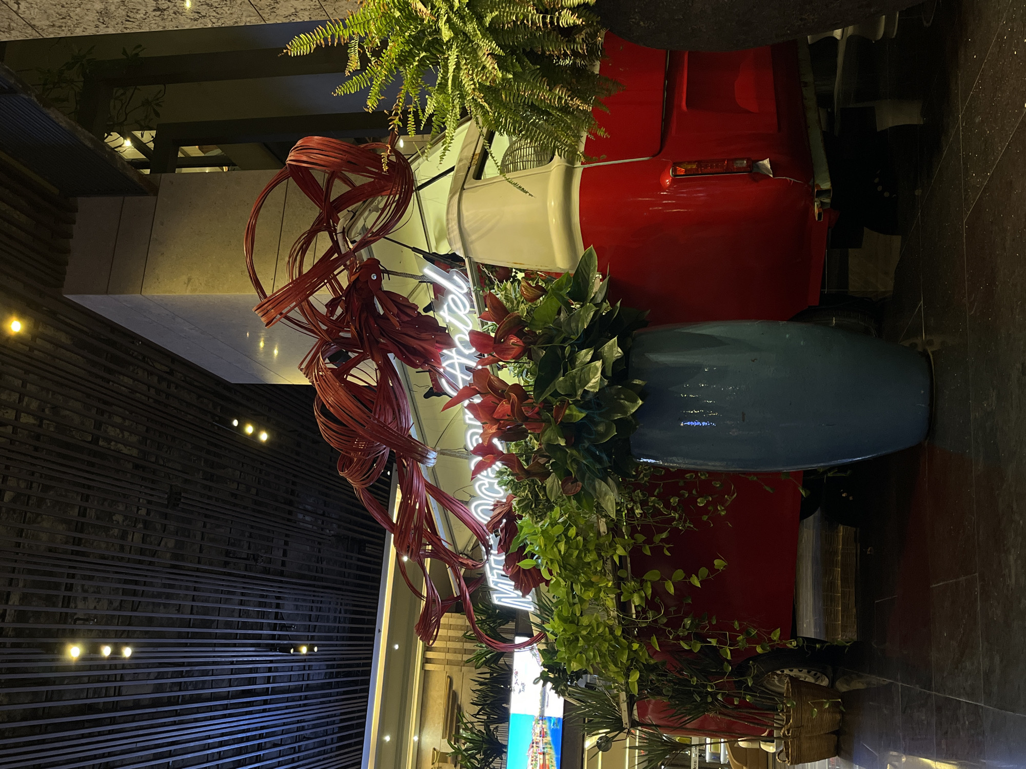 The image captures an indoor decorative setup in Đà Nẵng, Vietnam. A vintage red and white car is the central element. Ferns and other plants are arranged on and around the car, along with a blue vase. A red wicker-like decorative piece and a neon sign with the word "Hotel" in bright white are also present. The background includes a wall with horizontal metal bars and recessed lighting. The floor appears to be dark marble or tile.