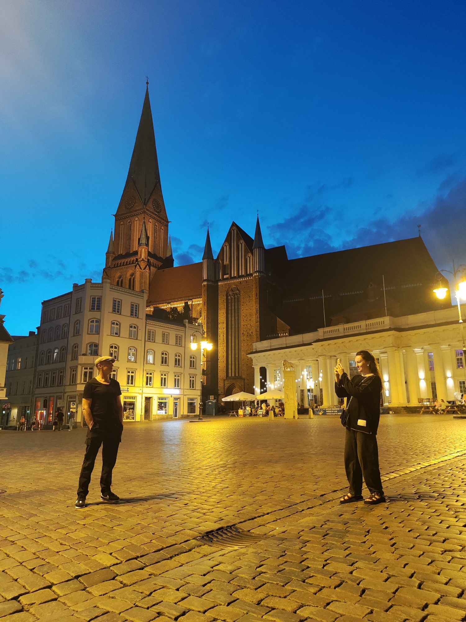 This image depicts the Domkirke (Schwerin Cathedral) in Schwerin, Germany, at dusk. The cathedral's tall spires and red-brick architecture dominate the background, illuminated by the fading natural light and artificial lighting from the surrounding buildings and streetlights. In the foreground, a cobblestone plaza is visible.
On the left side of the plaza, a person wearing a black t-shirt, black pants, and a cap stands with their hands in their pockets, looking towards the left. On the right side, another person, dressed in black clothing, is holding up a smartphone to take a photograph or video. This individual appears to be a woman with long hair.
The plaza itself is paved with cobblestones, and several streetlights cast a warm, yellow glow on the scene, creating long shadows. In the mid-ground, there are buildings with illuminated windows and signs, indicating shops or restaurants. There are also outdoor seating areas with people dining or socializing, as well as tables and chairs set up. The sky is a deep blue, transitioning into twilight, with some scattered clouds.