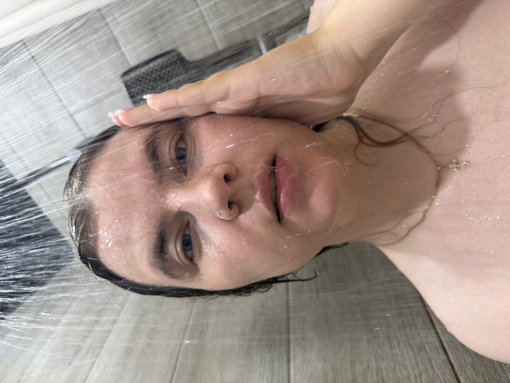 A woman is shown from a close-up, slightly angled perspective, presumably in a shower. Water streams across her face and body. She has fair skin, blue eyes, and dark eyebrows. A gold necklace with a small cross pendant is visible around her neck. She has a silver ring in her left nostril. Her hand is on her forehead. The background consists of shower tiles and shower fixtures. This image does not allow the exact location to be determined.