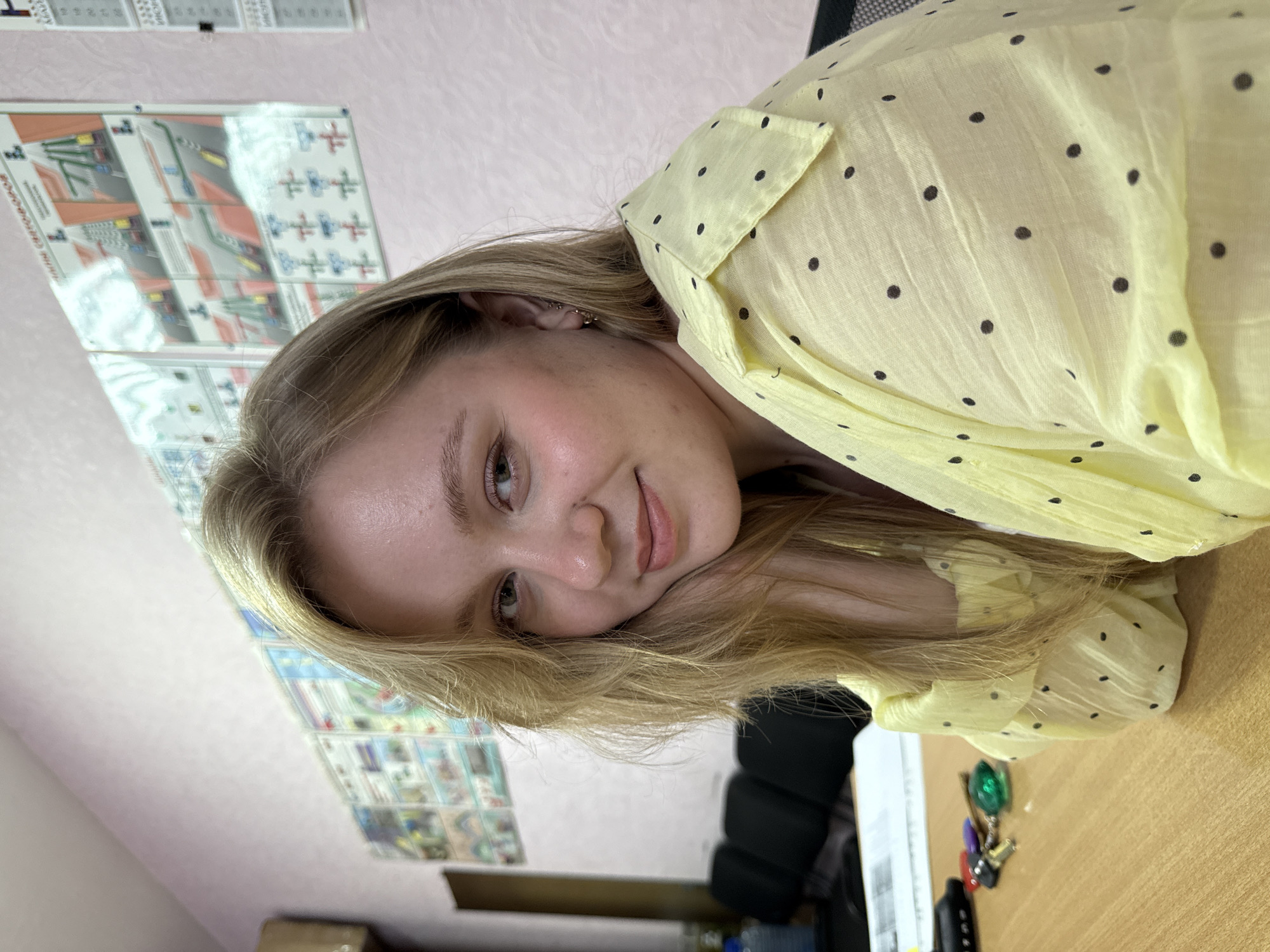 The media file is a close-up, head-and-shoulders shot of a young woman lying on her side, with her head propped up on her hands. She has blonde hair and appears to be looking towards the camera with a gentle smile. She is wearing a yellow blouse with black polka dots.
The background is blurred, but appears to be an indoor setting. There are some papers or posters on the wall behind her, with what look like diagrams and text, some of which are too blurry to read. On the surface in front of her, which is likely a desk, there are a few keys and a small green object. The overall impression is casual and relaxed, possibly at home or in an office. The lighting suggests it could be daytime, but there are no clear indicators of the time of day or weather. There is no visible text that can be clearly deciphered in the image. The context of Chaikovsky, Russia is noted but not directly evident from the visual cues.