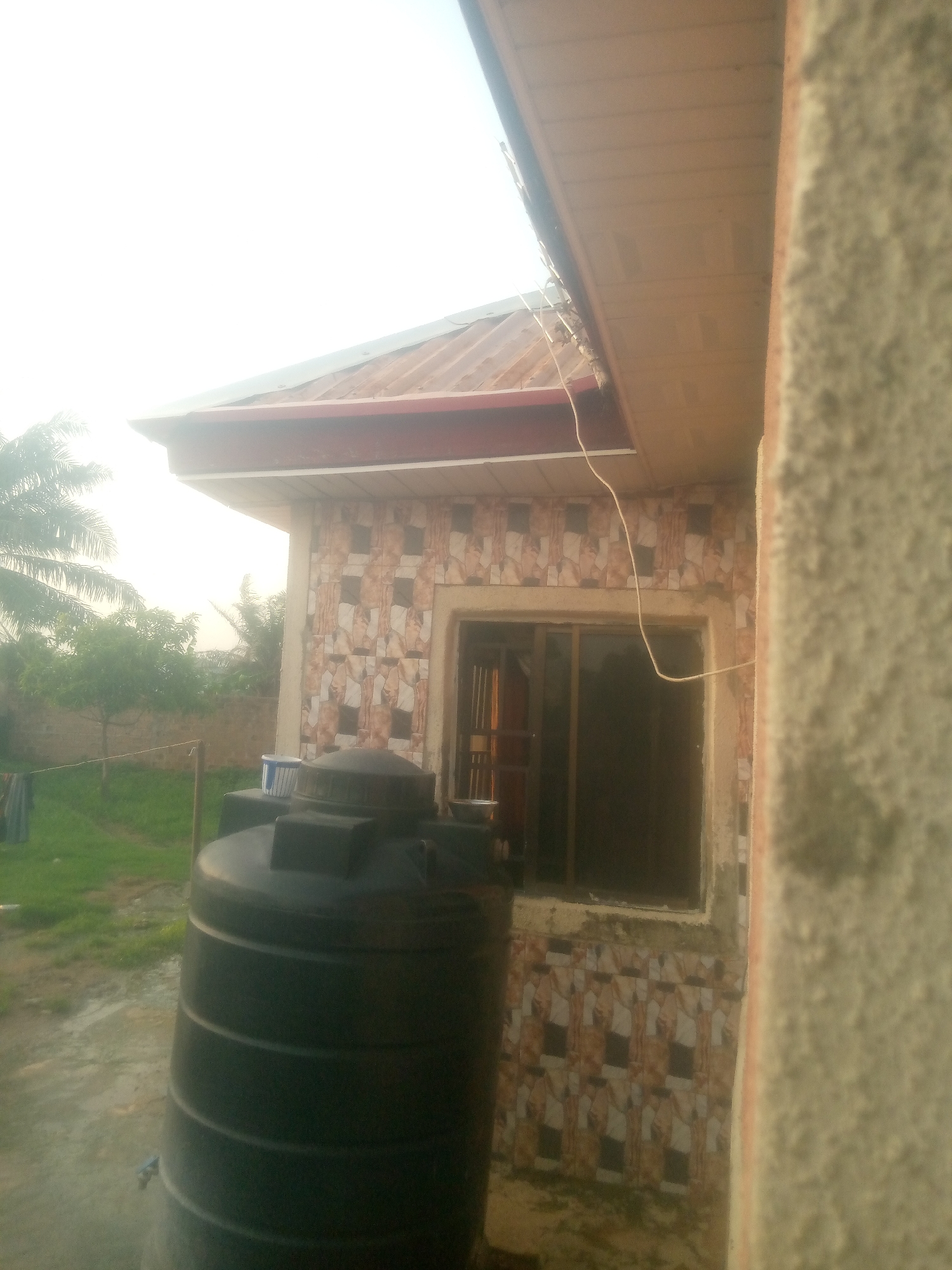 The image shows an exterior view of a building with a corrugated metal roof and a brown soffit. A large black water tank is positioned in the foreground, to the left of the frame. The building's wall is tiled with a pattern of beige, white, and dark brown squares, and there is a window with a dark frame. To the left of the building, there is a grassy area with some trees and laundry hanging on a line. The overall scene appears to be an outdoor space, possibly a residential compound. The location is identified as Jauro Yinu, Nigeria.