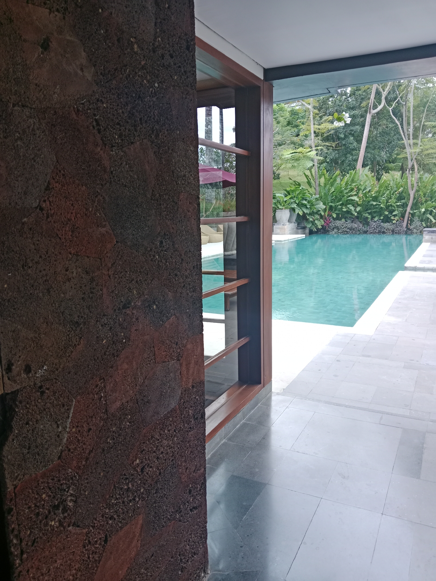 A close-up of a textured, dark red stone wall fills the left side of the frame. To the right, a wooden-framed opening reveals a bright turquoise swimming pool surrounded by lush green foliage and trees. The pool area is bordered by light-colored tiled flooring. The scene appears to be located in a tropical resort or private residence in Kuta Selatan, Indonesia, a region known for its beautiful beaches and upscale accommodations.