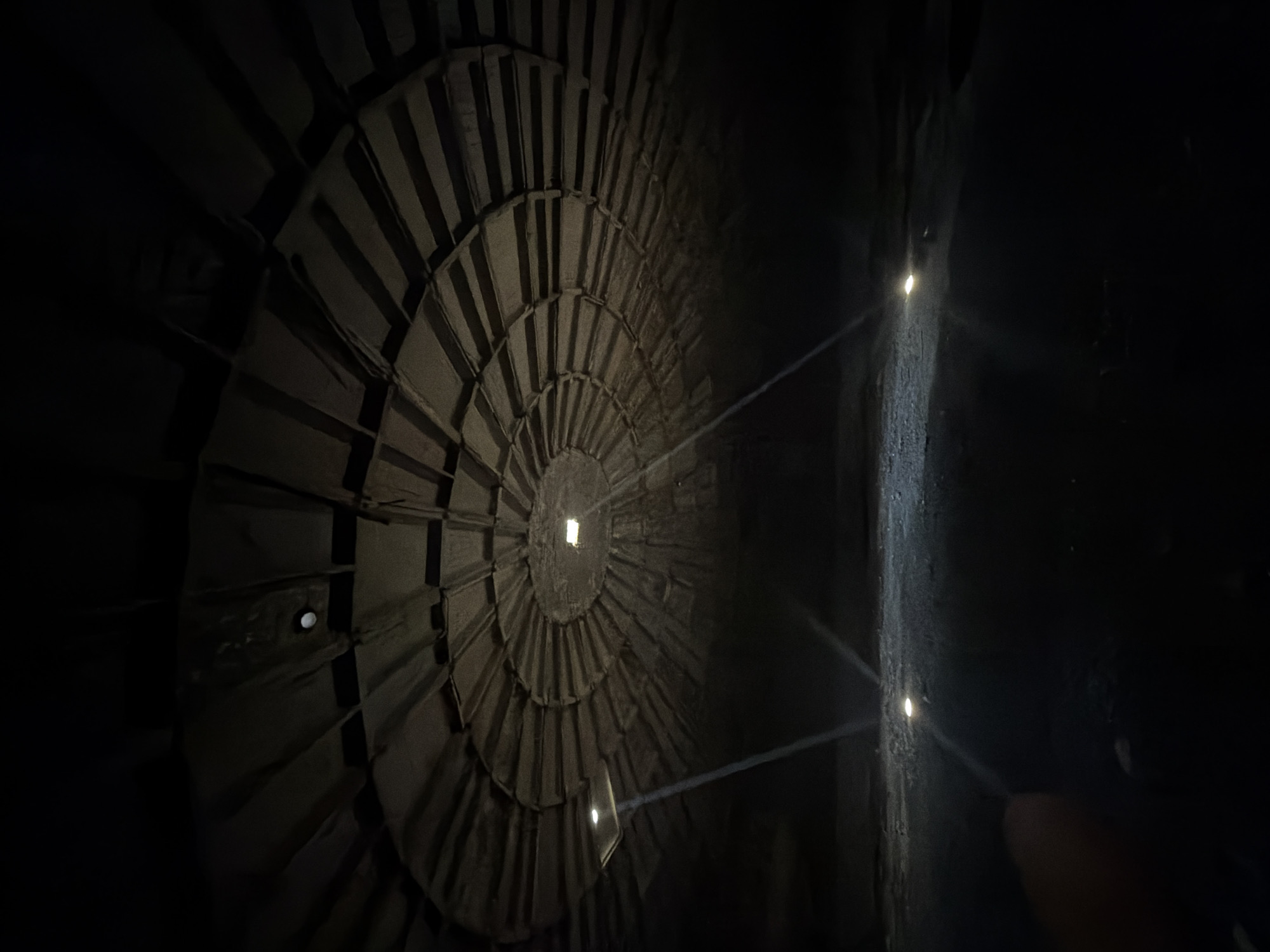 The image captures the interior of the Tbilisi History Museum (also known as the Tbilisi History Museum Karvasla) in Tbilisi, Georgia. The view is of a large circular architectural feature resembling tiered steps or a turbine-like structure, constructed from a dark-colored material. A small square opening is visible at the center of the circular structure. Light enters through this opening and other small holes in the structure.
To the right, a rough, dark wall is illuminated by three small, bright points of light. Light beams emanate from these points, creating a contrasting linear pattern against the curve of the circular structure.
The overall environment is dark, with limited illumination highlighting the texture and form of the interior architecture. The scene is still and appears unoccupied.