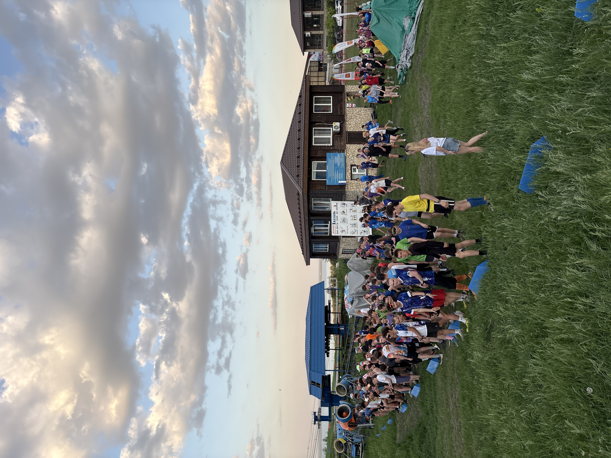 A large group of people stands on a grassy field in front of a building. The building has multiple stories and a brown roof. Behind the building are more people. A blue structure stands near the front of the crowd. The sky is cloudy with some blue patches visible. The location is Khvalynsk, Russia.