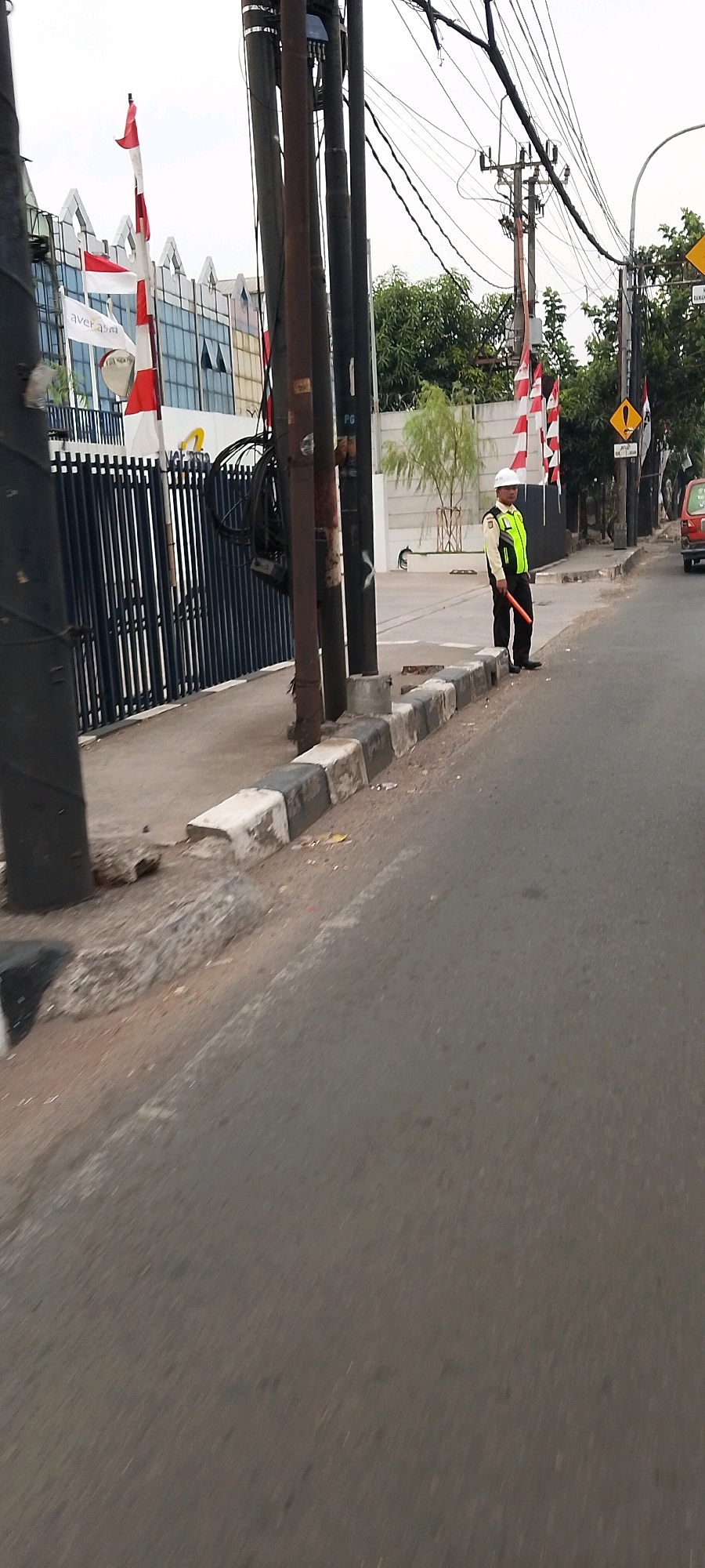 The image depicts a street scene in Bekasi, Indonesia, with a focus on a traffic director standing on the sidewalk. The traffic director is wearing a white hard hat and a high-visibility lime green vest over a light-colored shirt and dark pants. They are holding a red traffic baton. The traffic director stands near a row of black metal gates, which lead to a building with a modern architectural design featuring angular white elements and large windows. Several Indonesian flags, red and white, are visible on flagpoles near the entrance of the building.
Behind the traffic director and the gates, a concrete wall separates the property from the street. A thin, deciduous tree with light green foliage grows behind the wall. Electrical poles and a complex network of overhead wires dominate the mid-ground and background on the right side of the image. Traffic signs are mounted on poles; one yellow sign with a black exclamation mark is clearly visible. The street itself is paved with asphalt and shows some wear. The curb separating the sidewalk from the road is made of alternating black and white concrete blocks, some of which appear damaged. The image appears to have been taken from a moving vehicle due to the slight motion blur, particularly in the foreground.