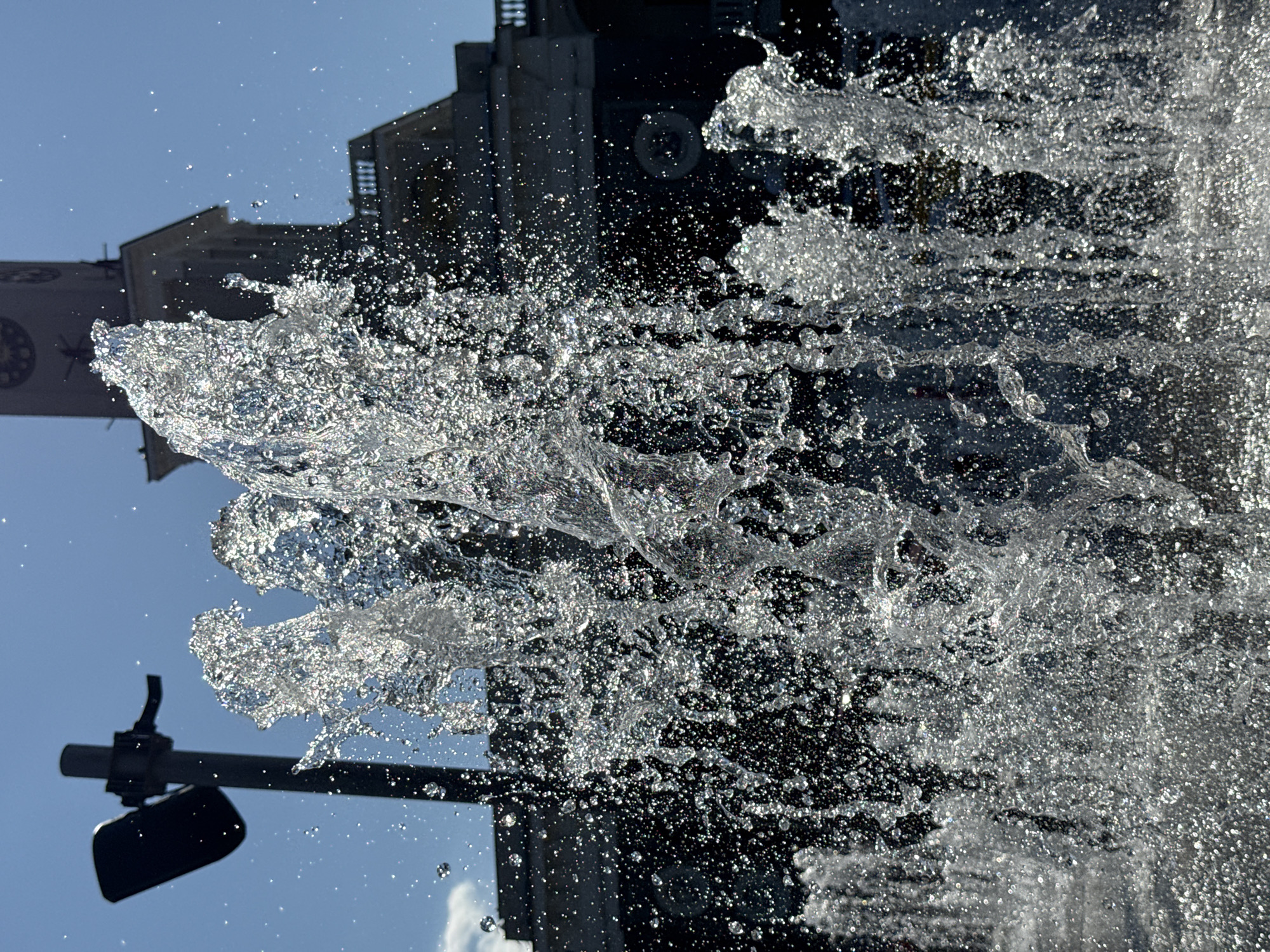 This is a close-up, low-angle, outdoor photograph of a fountain spray against a bright blue sky. The focus is on the dynamic, fractured streams of water, catching the sunlight and creating a shimmering effect with many small droplets. In the background, a dark, somewhat blurred structure with architectural details, including a clock tower, is visible. To the left, a dark object on a pole, possibly a light fixture, is also present. The scene appears to be in a public urban space.