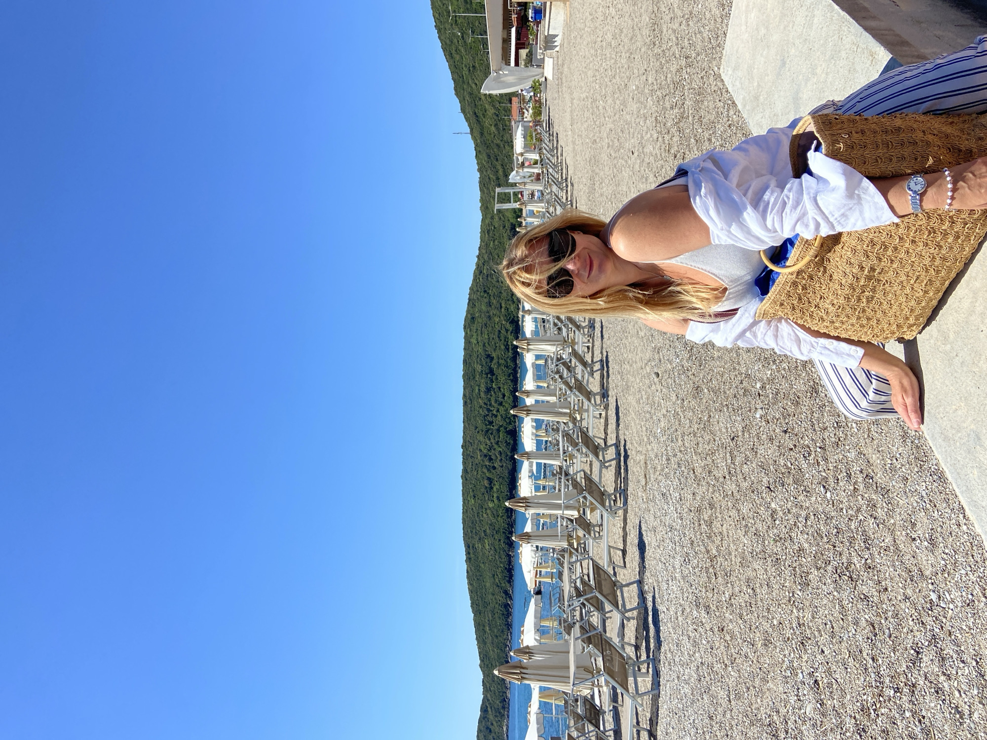 A woman with long blonde hair and sunglasses is sitting on a stone bench at a beach in Prijevor, Montenegro. She is wearing a white shirt over a grey tank top and blue and white striped pants, and has a straw bag next to her. The beach has rows of sunbeds with white umbrellas and is made of small pebbles. Behind the beach, there is a dense green forest. The sky is clear and blue.