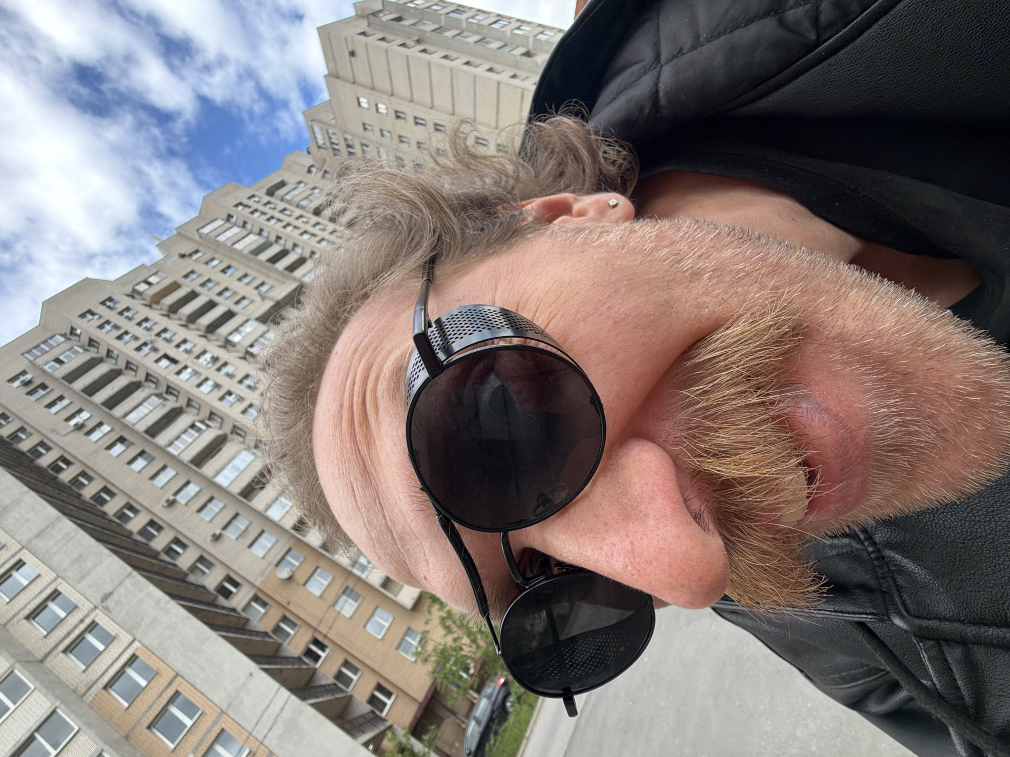 The photograph presents a close-up, low-angle perspective of a person wearing black, round sunglasses, positioned against a backdrop of an apartment building and sky in Saint Petersburg, Russia. The individual has blond facial hair and a visible ear piercing. The building is a high-rise apartment block with rows of windows, a standard architectural style of the city. The sky is partially visible with light cloud cover. The person is wearing a black jacket or coat.