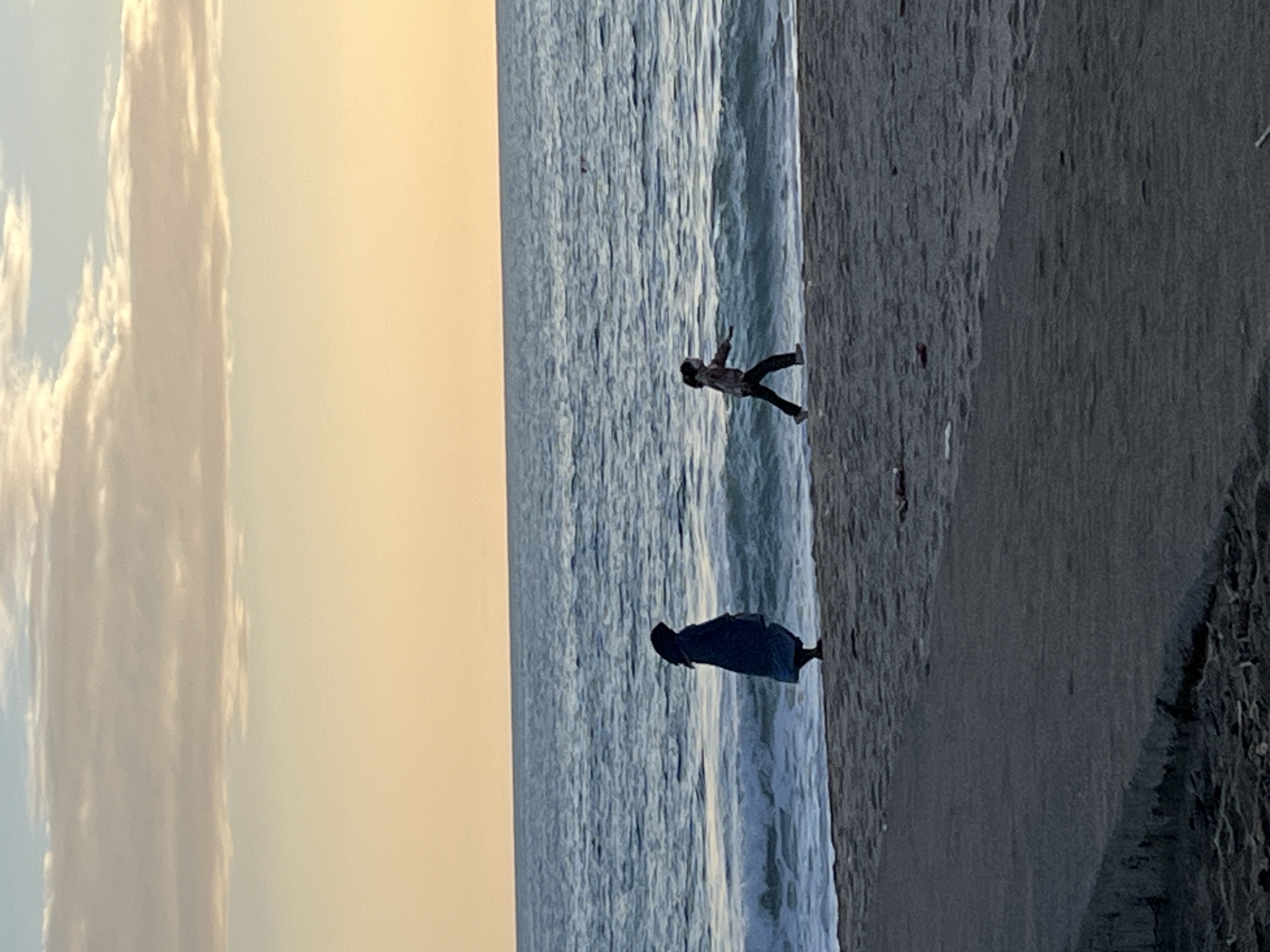 A landscape photograph shows a beach scene in Alanya, Turkey. The beach occupies the right side of the frame and extends to the bottom. Adjacent to the beach is the sea with visible waves. Two individuals are present: one is near the waves, appearing to walk or stand, dressed in a blue robe. The second figure is further out in the water, positioned in a way that suggests the person is either falling or jumping. To the left is the sky with some cloud cover that is in a gradient color with darker colors at the top and lighter colors at the bottom of the frame. The photo suggests either early morning or late evening due to the lighting conditions.