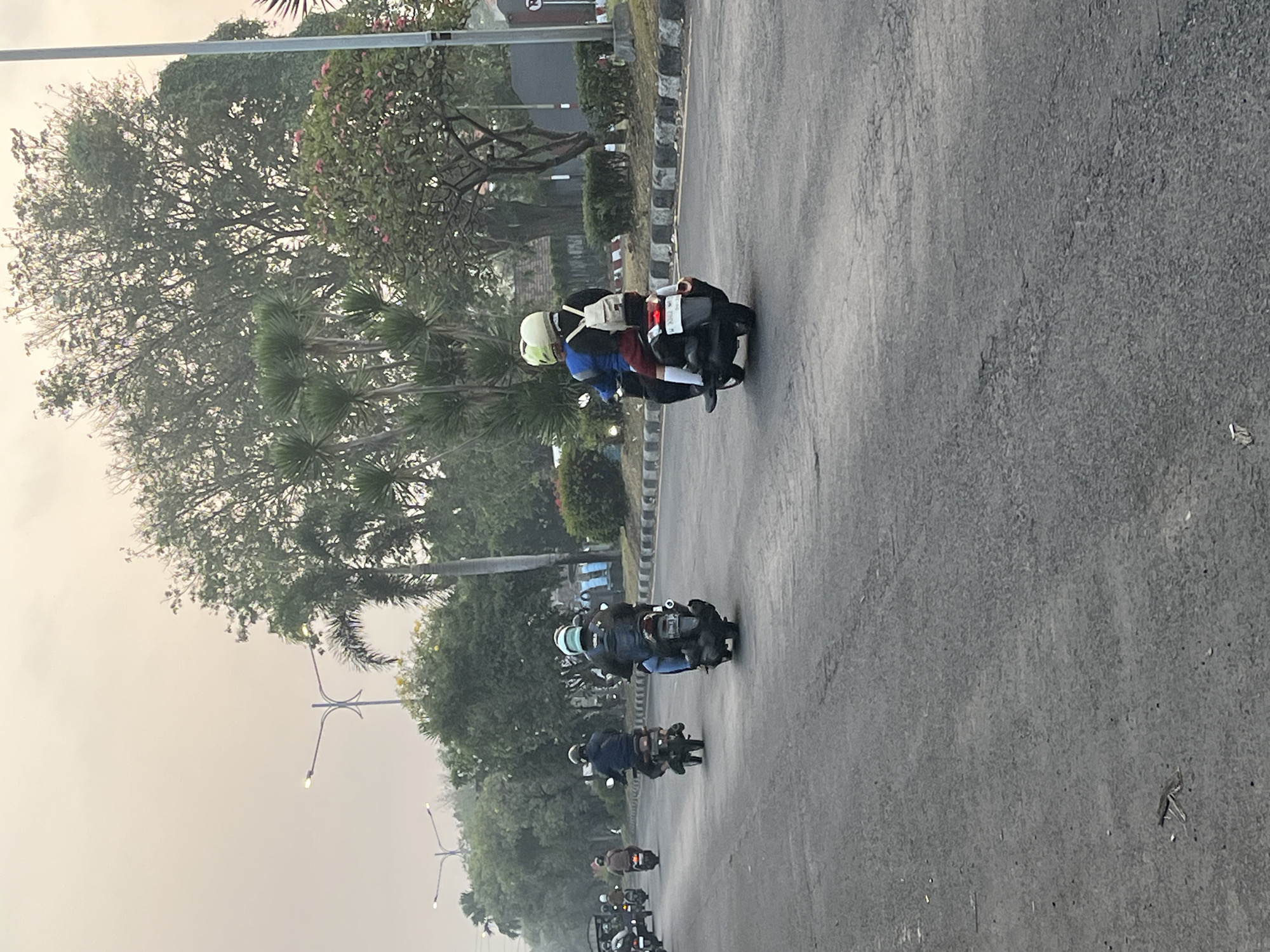 The image depicts a street scene in Kludan, Indonesia, characterized by overcast skies and lush greenery lining the road. Several motorcycles are traveling on the paved road, some carrying multiple passengers. In the foreground, a motorcycle is ridden by two individuals wearing helmets, the passenger in the back has a white backpack. Further down the road, other motorcycles are visible, with riders also wearing helmets. The environment includes trees, palm trees, and bushes, with a streetlight and utility pole visible in the background. The road has distinct white lines marking lanes and a concrete barrier with decorative elements along the side. The overall impression is of a typical day with local transportation on a suburban or semi-urban road.