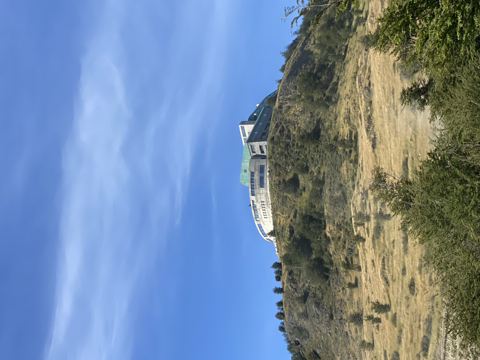 The image captures a scenic view of a large building nestled into a hillside, set against a clear blue sky.
The upper portion of the frame is dominated by a bright, cerulean blue sky, sparsely dotted with thin, wispy white clouds that stretch across the expanse. The daylight appears clear and bright, suggesting a sunny day.
Towards the middle and lower-right of the frame, a multi-story building is prominent, seemingly built into the natural contour of a steep hill or mountain slope. The architecture of the building features modern elements, with sections appearing to be constructed from grey or stone-colored materials. A distinctive feature is a large, possibly oxidized copper or similar material, green-colored roof on a central section of the building. Below this, there's a curved, white facade with multiple windows, possibly balconies or a panoramic viewing area, suggesting it might be a hotel or resort.
The building is surrounded by and integrated into the natural landscape of the hillside. The slope is covered with a mix of golden-brown dry grass, indicative of a dry climate or season, interspersed with darker green patches of scrub brush and scattered conifer trees. The terrain appears rugged and uneven, with rocks and variations in elevation. More dense foliage and trees are visible on the far right side of the image, framing the view.
There are no people or discernible activities taking place in the image, which presents a serene and expansive landscape with a significant architectural presence. No text is visible in the image.