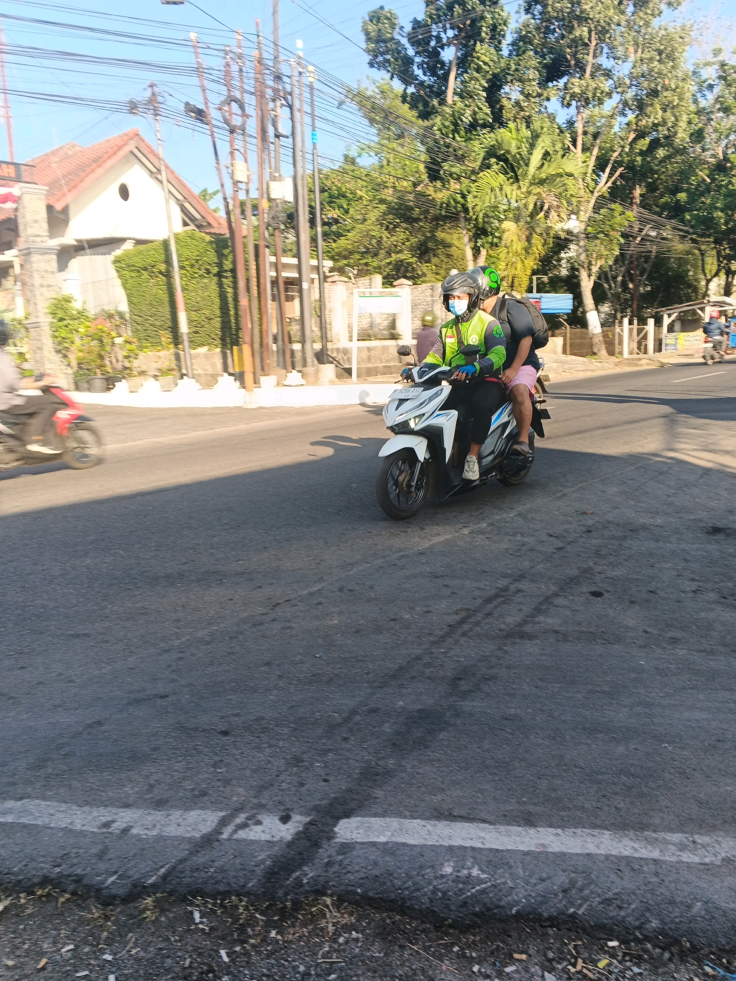 Two individuals are riding a white motorcycle on a paved road in Semarang, Indonesia. The driver, wearing a green jacket and a helmet, is operating the motorcycle. The passenger, also wearing a helmet, is seated behind the driver. The road is bordered by trees and buildings. The scene is illuminated by natural daylight.