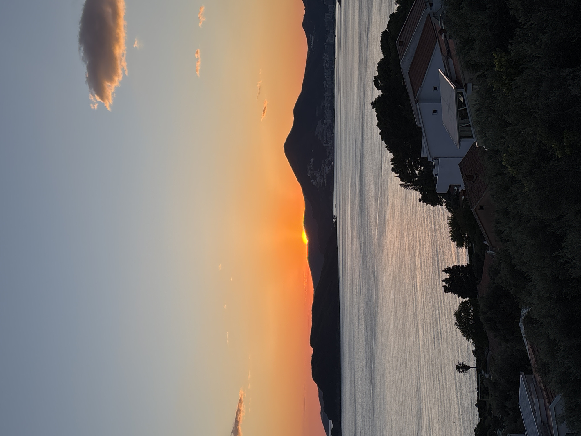 The image shows a sunset over water. The sun is partially obscured by a mountain range, casting an orange glow across the sky. To the right, there are buildings and trees lining the water's edge. The buildings are white with red tile roofs. The location is Unknown, Unknown.