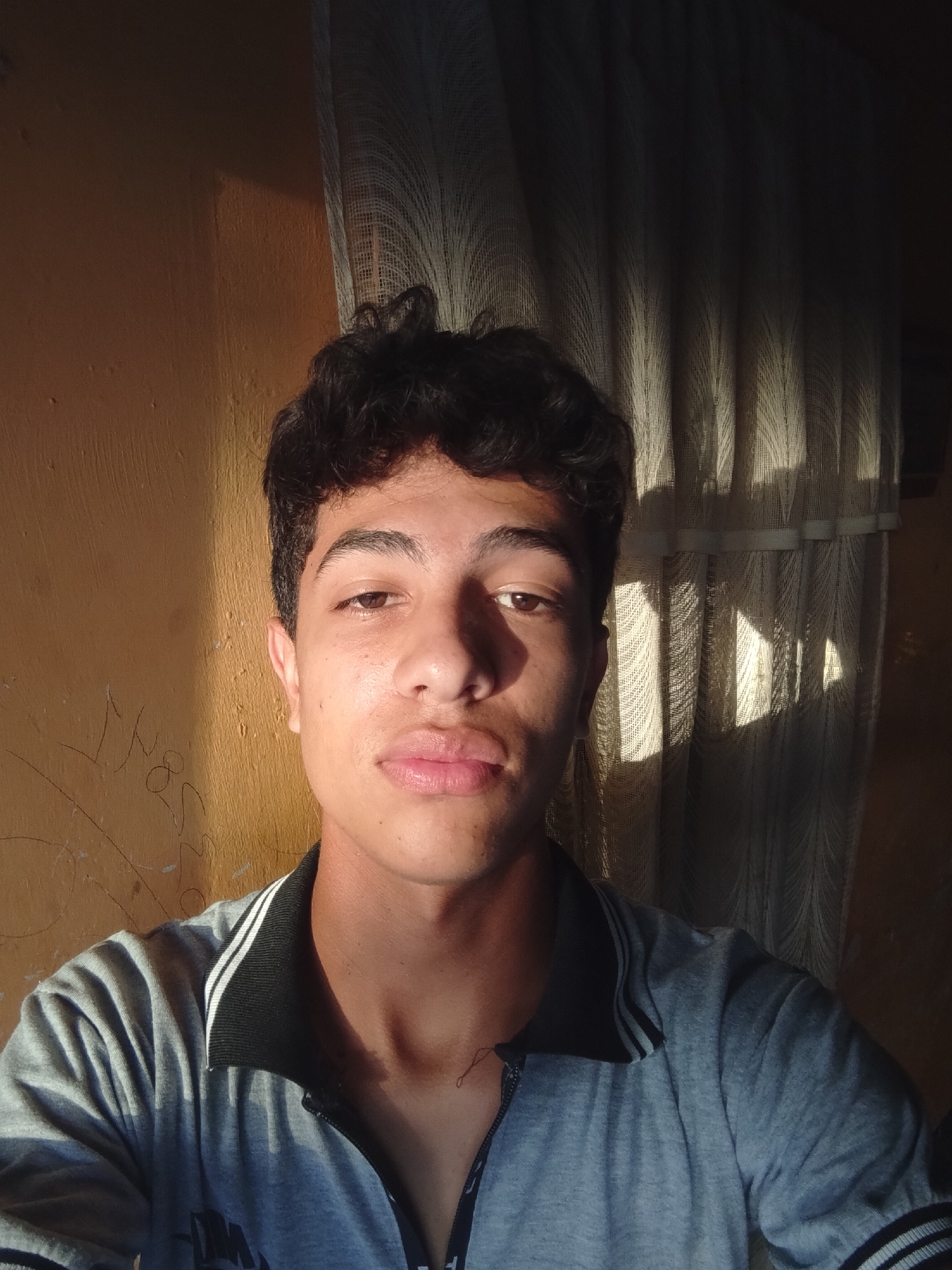 The image is a selfie taken indoors, likely in Santa Ana, Venezuela, given the context of the prompt. A young male with dark, curly hair is centered in the frame, looking directly at the camera. He is wearing a grey polo shirt with a black collar and white stripes. The background consists of a textured, yellowish wall on the left, with some faint graffiti marks. On the right, a light-colored, patterned curtain is visible, with sunlight casting vertical shadows across it. The lighting suggests the sun is low in the sky, casting warm light on the left side of the subject's face and the wall, while the right side is in shadow. The overall impression is of a casual, personal moment captured in natural light.