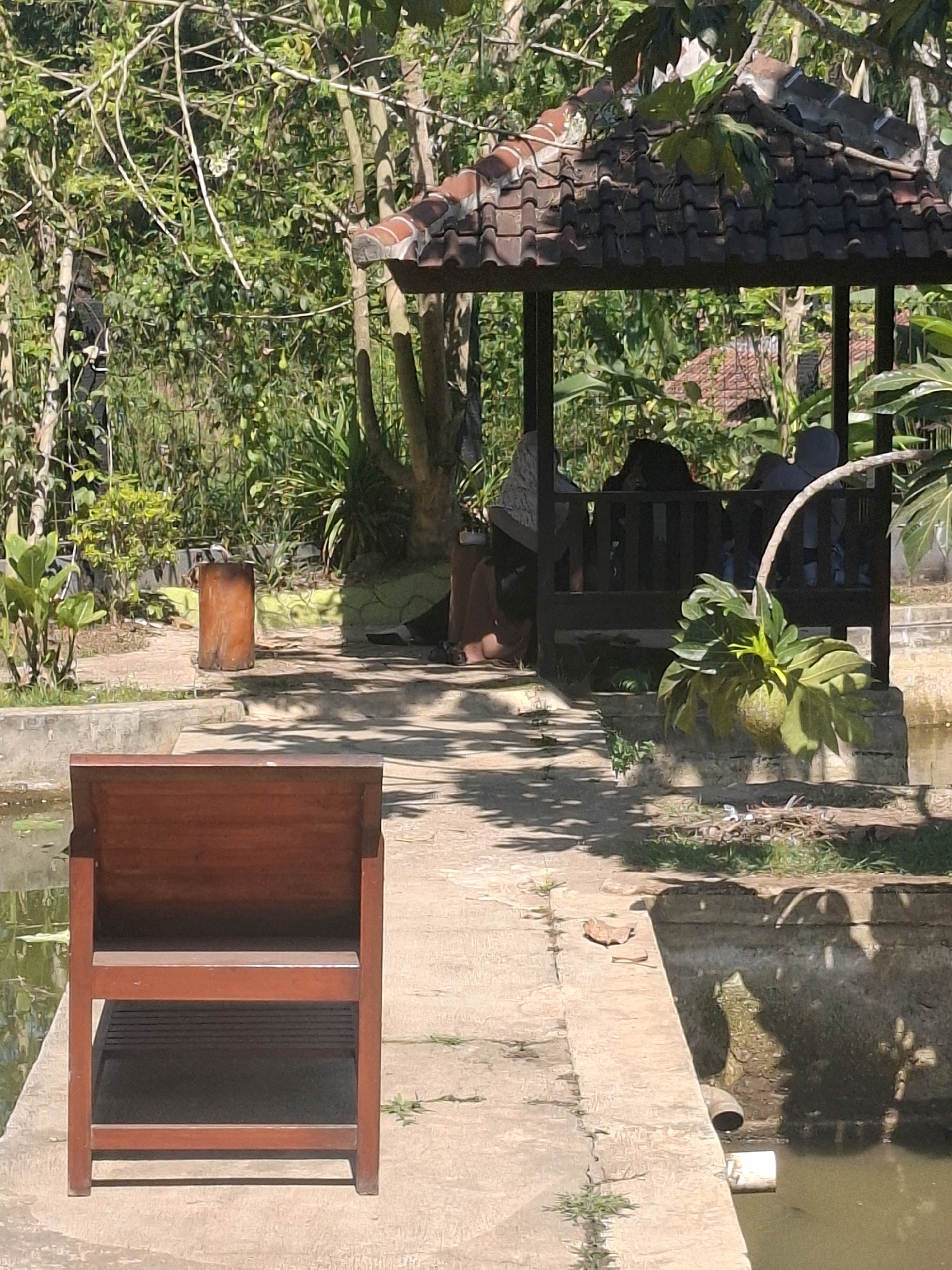 In Cibentang, Indonesia, a sun-drenched outdoor scene unfolds. A rustic wooden chair stands prominently in the foreground, facing a pathway that leads towards a shaded gazebo. Within the gazebo, several individuals are seated, their figures partially obscured by the dim lighting and foliage. The gazebo itself is adorned with a traditional tiled roof. Surrounding the gazebo and pathway is abundant greenery, with trees and various plants creating a lush, natural environment. A body of water, possibly a pond or canal, is visible to the left and right of the pathway, reflecting the sunlight and casting shadows. A cylindrical object, perhaps a bin or planter, rests on the ground to the left, near the pathway. The overall atmosphere is serene and suggests a place of relaxation and quiet contemplation.
