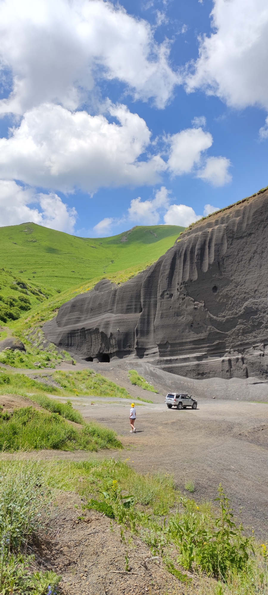 A person wearing a yellow hard hat and a white t-shirt stands in a rocky landscape. The person is facing away from the camera, looking towards a dark, layered rock formation with openings that appear to be caves. A silver SUV is parked to the right of the person, on a gravelly surface. Rolling green hills are visible in the background, under a blue sky with white, puffy clouds. The scene suggests a daytime setting, likely during fair weather, given the clear sky and bright lighting. The location appears to be a natural geological area, possibly in Fantan, Armenia, characterized by its dramatic rock formations and verdant hills. The overall mood is one of exploration or scenic appreciation. No visible text is present in the image.