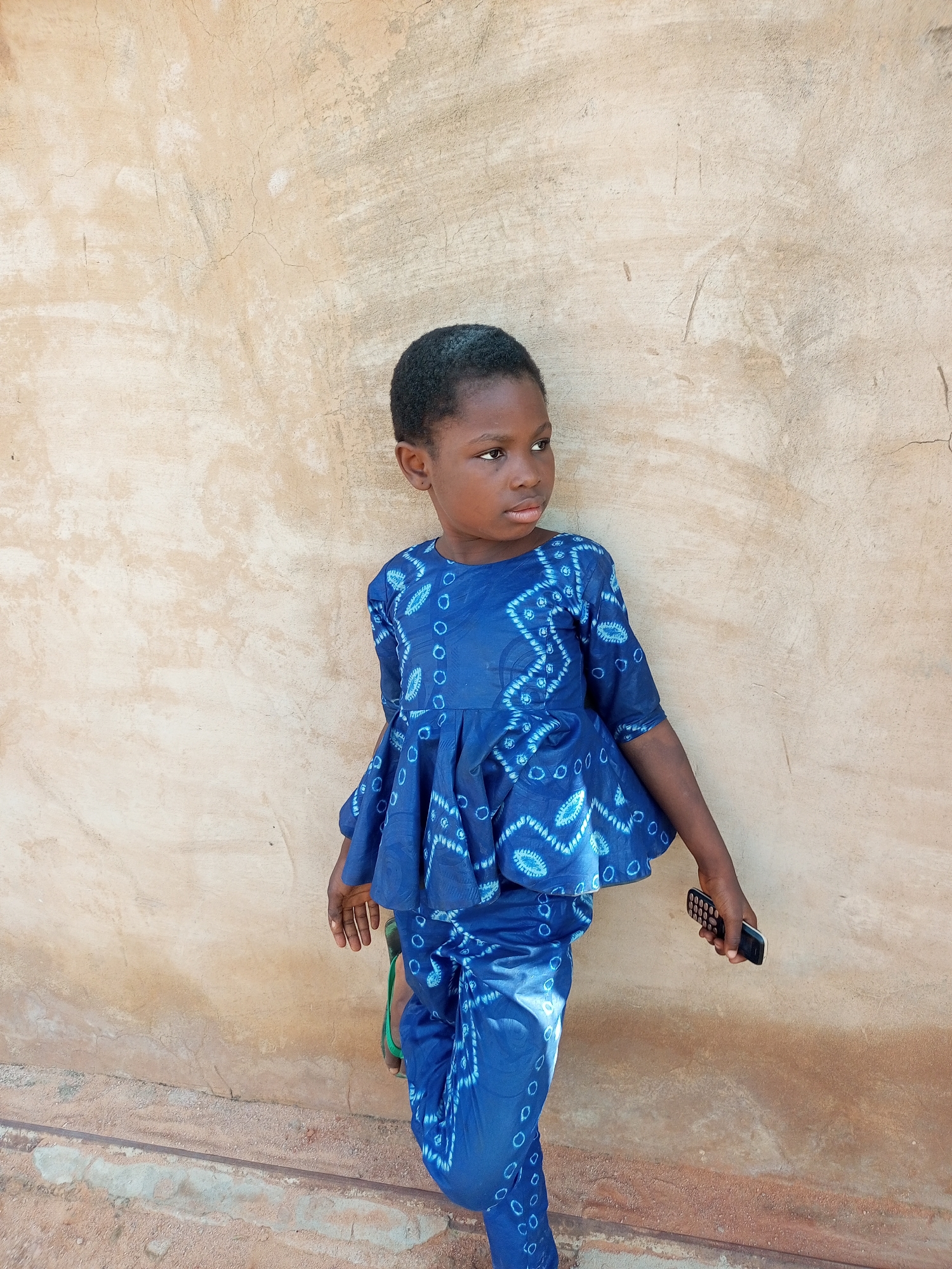 A young Black girl stands against a textured, pale beige wall, her body angled to her right. She is wearing a blue, tie-dye print outfit consisting of a peplum top and matching trousers. Her short, dark hair frames her face. She is looking to her right, with her gaze slightly upward. Her right hand holds a black mobile phone. Her left arm is bent at the elbow, with her hand resting near her hip. She is wearing green flip-flops, only partially visible beneath her trousers. The wall has a rough, weathered appearance with visible cracks and some discoloration. The ground in front of the wall is a mix of dirt and small stones, with a raised concrete or cement edge. The overall scene appears to be outdoors, possibly in a courtyard or alleyway in Tempure, Nigeria.