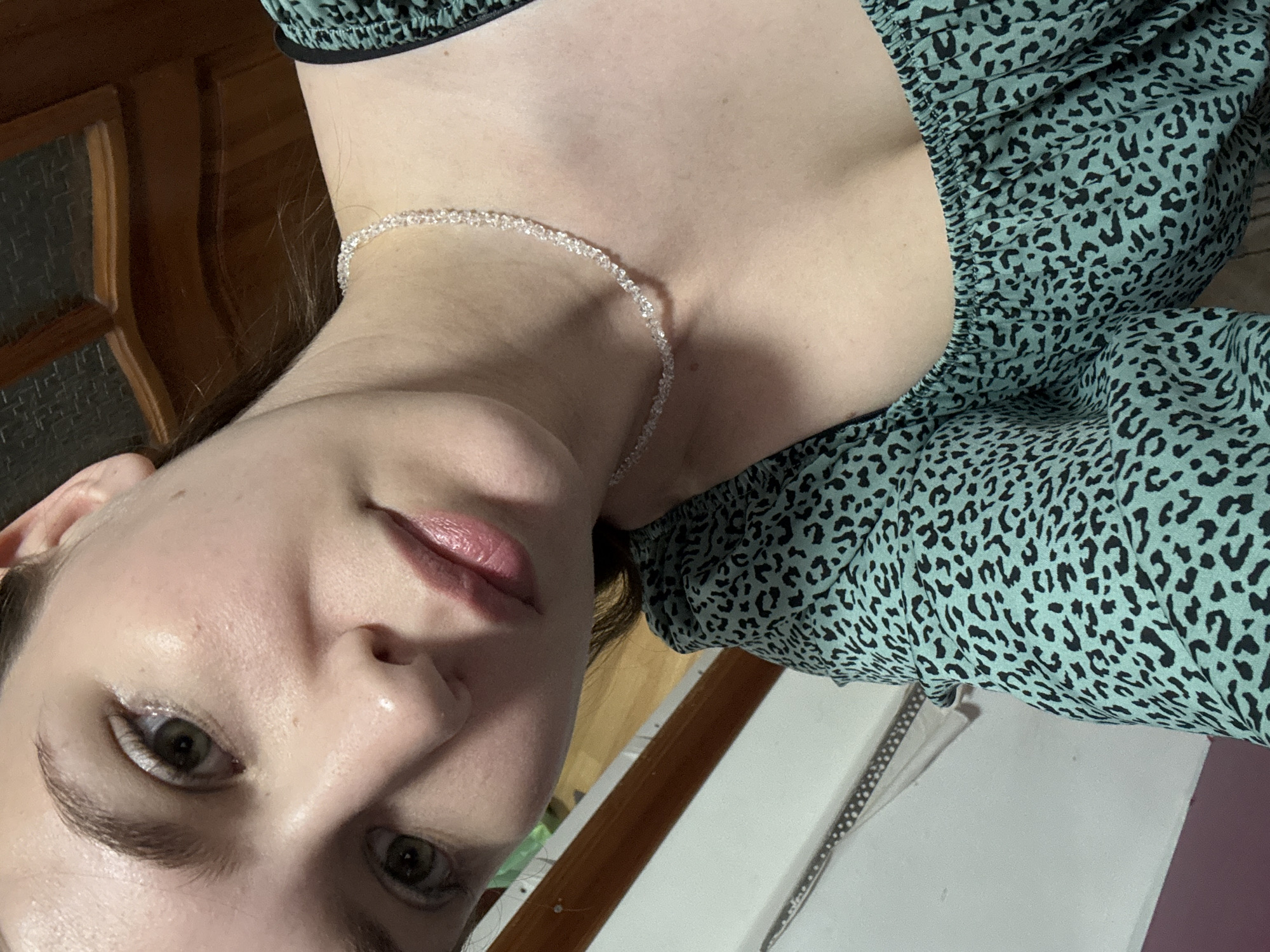 The image shows a close-up, angled selfie of a young woman with fair skin and light green eyes. She is wearing a teal and black leopard print top with a square neckline and a delicate, clear beaded necklace. Her lips are slightly parted and appear to be wearing a subtle pink lipstick. Her skin has a dewy finish, and she has natural-looking eyebrows and subtle eye makeup. In the background, a wooden door with dark wood paneling is visible. The overall lighting suggests an indoor setting, possibly a home. The image does not provide enough information to determine its specific location within Chaikovsky, Russia.