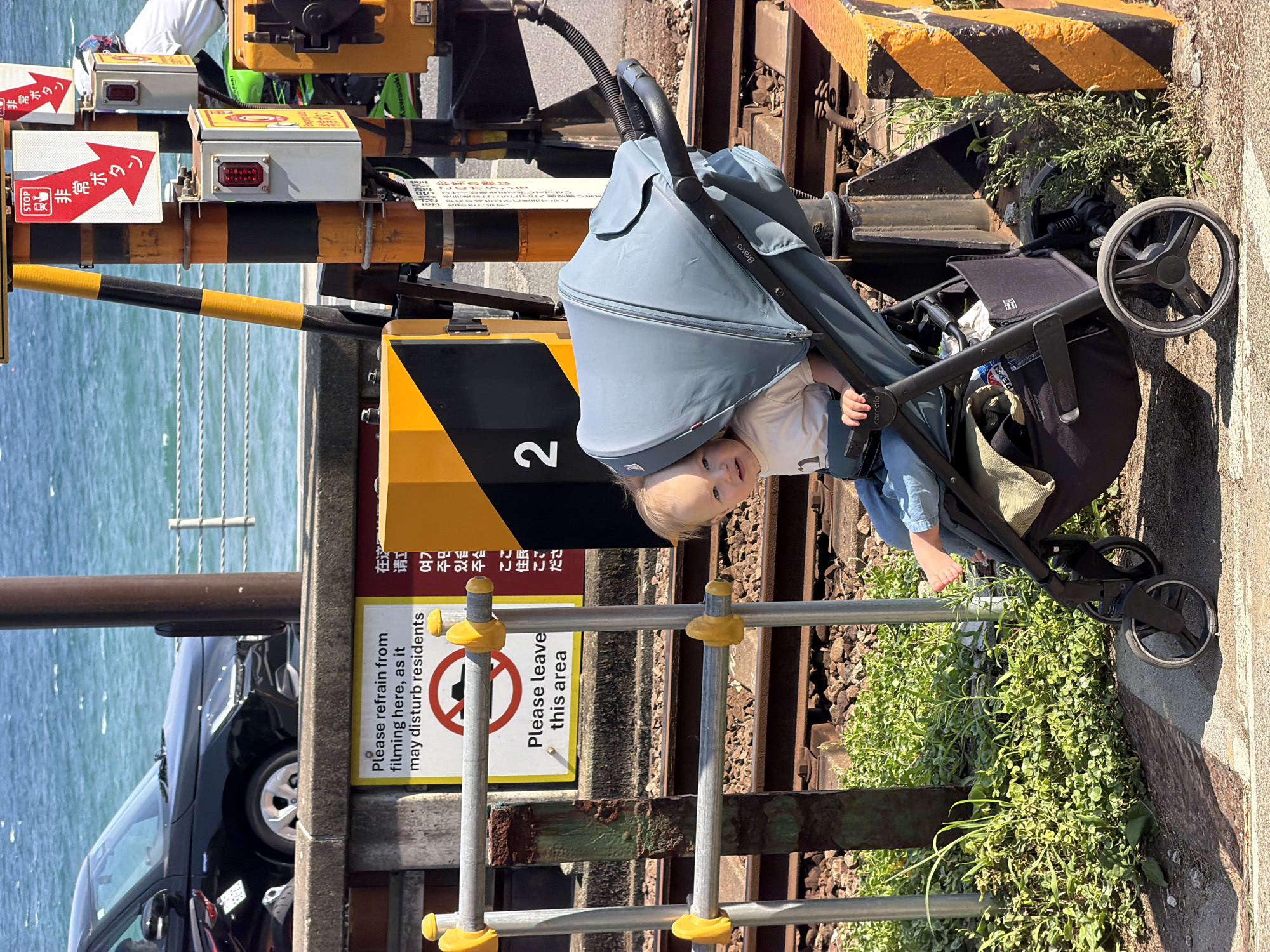 A toddler sits in a blue stroller next to train tracks in Kamakura, Japan. The child, wearing a white t-shirt and blue jeans, looks towards the camera. The stroller is positioned on a paved walkway adjacent to the tracks. A yellow and black striped barrier is visible to the right of the stroller. In the background, the ocean is visible, with a black car parked on a road overlooking the water. A sign with Japanese text and a "STOP" symbol is attached to a pole near the train tracks. The scene appears to be a sunny day.