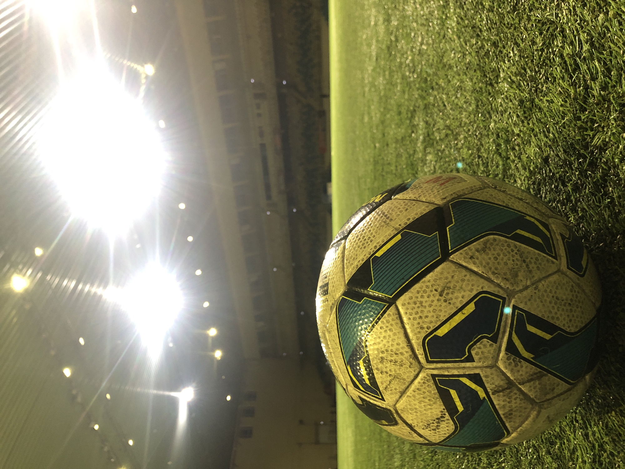 The image shows a soccer ball on artificial turf, likely a soccer field. The ball is mostly white with dark blue and yellow accents. Bright stadium lights are visible in the background, suggesting it is nighttime. The exact location in Krasnoyarsk, Russia, cannot be determined from the image alone. There are no people visible.
