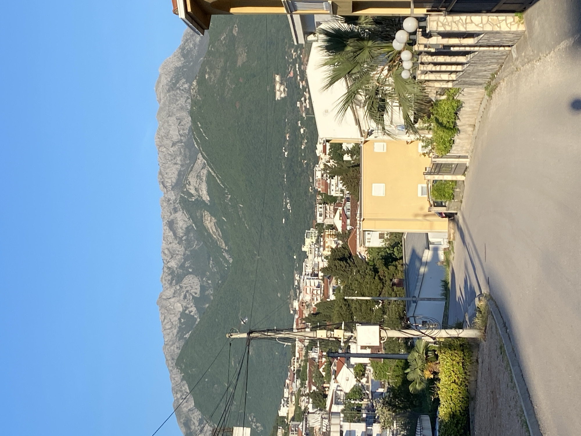 This image captures a scenic view of a town in Šušanj, Montenegro, situated between a rugged mountain range and, presumably, the coast, under a clear, bright blue sky. The perspective is slightly elevated, offering a sweeping look over the urban landscape and natural surroundings.
On the left side of the frame, a grand, rocky mountain range dominates, characterized by its grey limestone peaks and ridges. The lower sections of the mountain are densely covered in vibrant green vegetation, providing a natural transition to the developed area below. A few small, light-colored buildings are visible high on the verdant slopes, hinting at remote residences or structures.
The mid-ground and right side of the image reveal the town itself, densely packed with numerous light-colored buildings. Most structures feature white or light yellow facades complemented by distinctive red or orange tiled roofs, typical of the Adriatic region. These buildings are interspersed with lush green foliage, including several prominent palm trees and various bushes, creating a verdant urban environment.
In the immediate foreground on the right, a paved road or street is visible, gently curving through the landscape. Alongside this road, a decorative stone staircase with multiple spherical white lampposts ascends, suggesting access to higher terraces or buildings. A notable utility pole, heavily laden with a network of electrical wires, stands centrally in the foreground, adding an element of infrastructure to the otherwise natural and architectural scene.
The lighting indicates either early morning or late afternoon, as long shadows are cast by the buildings and utility poles, suggesting the sun is low in the sky. The weather is clear and sunny with no clouds visible. No people, animals, or discernible text are present in the image, conveying a sense of tranquility and an unspoiled natural setting alongside human development.