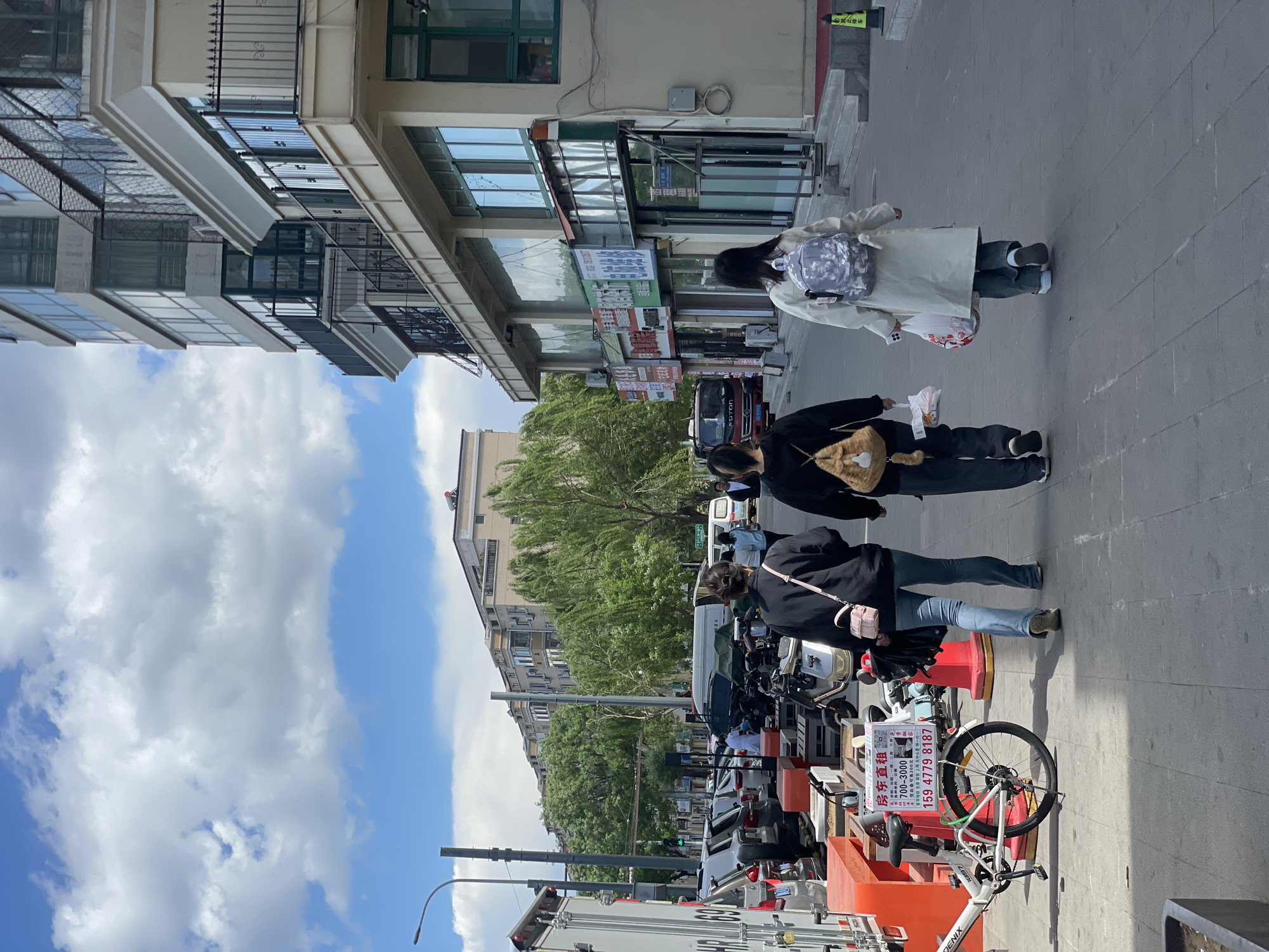 The image shows a street scene in what appears to be Changping District, China. Three people are walking on the sidewalk. The first pedestrian is wearing a light-colored coat with a backpack. The next pedestrian is wearing a black coat with a shoulder bag. The third pedestrian is wearing a black jacket, light denim pants, and is carrying a pink purse. On the right, an overturned bicycle is visible next to a traffic cone and a sign in Chinese characters advertising rental properties. Behind this is some street furniture and parked electric motorbikes. The street is lined with buildings of varying heights, with shops and residential units visible. The sky is blue with scattered clouds.