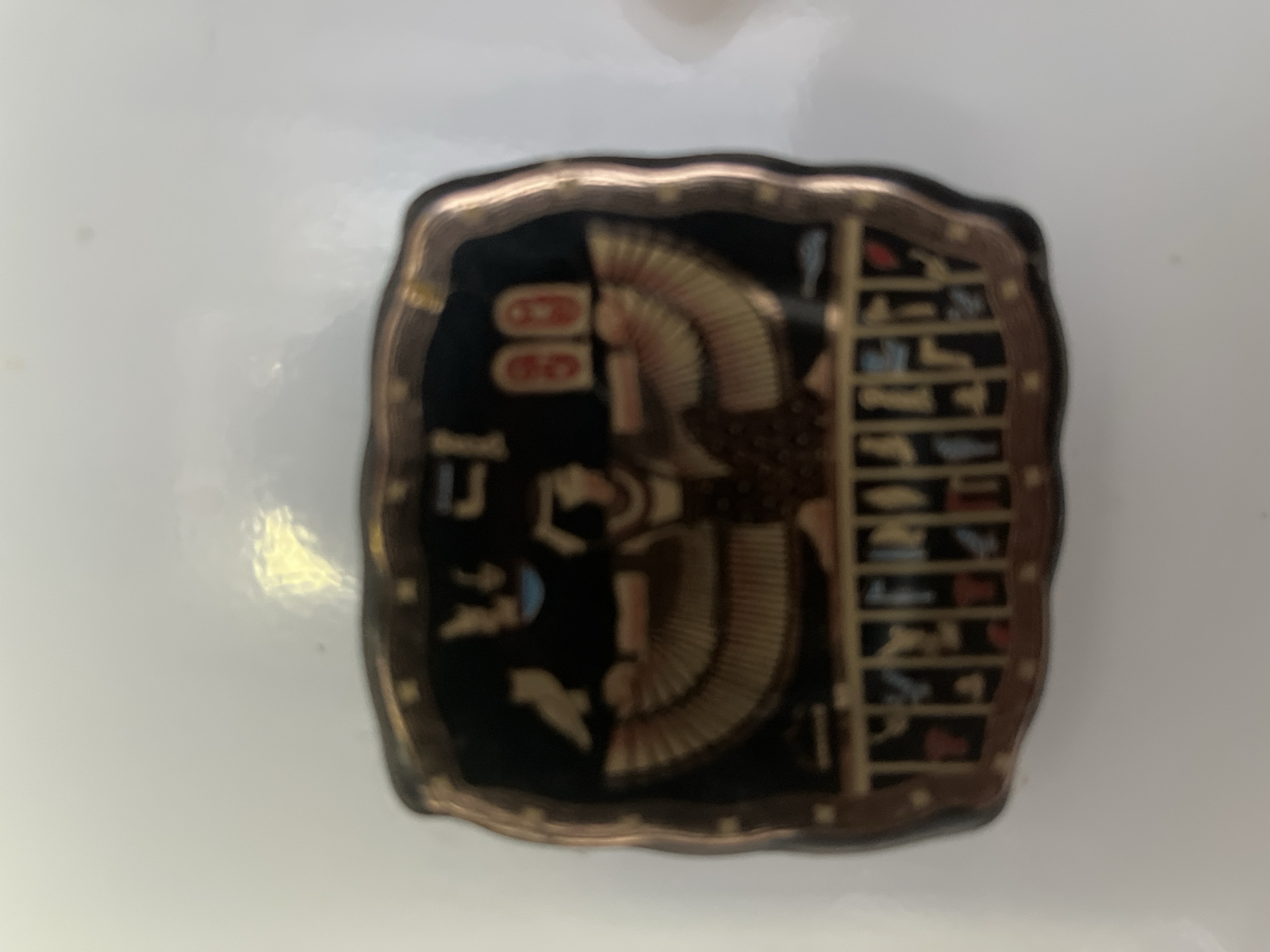 The media file showcases a dark, square-shaped decorative object with an ancient Egyptian theme, resting on a plain, light-colored surface. The object has distinctive scalloped or wavy edges, framed by a metallic gold or bronze border.
At the center of the object is a stylized depiction of a winged figure, likely a goddess such as Isis or Nephthys, facing left. The figure has a human-like torso and head, which wears an indistinct headdress. Large, elaborate wings spread symmetrically behind the figure, rendered in gold or bronze with a textured, feathered appearance.
The background of the object is predominantly black, providing a stark contrast to the metallic golden details. To the right of the central winged figure, several vertical columns of ancient Egyptian hieroglyphs are visible. These small symbols are rendered in gold or bronze and include various pictorial representations like birds, geometric forms, and other abstract shapes. On the left side of the object, a few larger, less structured hieroglyphic symbols are present, one of which resembles a shen ring or cartouche.
The object appears to be a decorative plaque or souvenir, possibly crafted from resin or a similar material with metallic accents. There is no activity or event depicted beyond the static image of the object itself. The surrounding area is an out-of-focus, uniform white or light grey surface, likely a table or countertop. No specific time of day or weather conditions are discernible from the image. While the location context is Minsk, Belarus, the object itself features iconography from ancient Egypt.