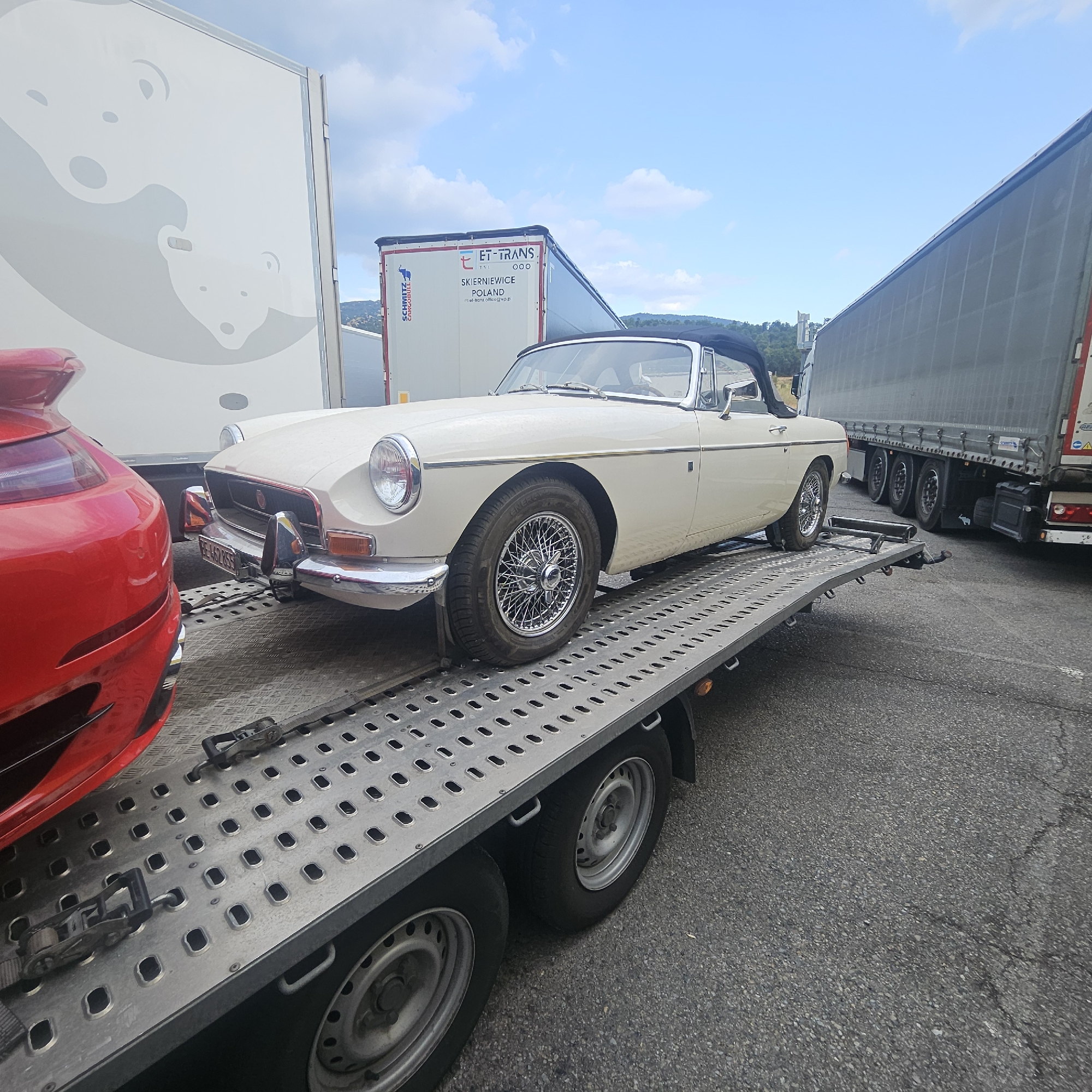 The image shows a light-colored classic convertible on a car transporter trailer, positioned on an asphalt road. To the left, a portion of a red sports car is visible. In the background, several large semi-trailer trucks are parked, one with the branding "ET-TRANS POLAND". The sky is blue with scattered clouds. The scene appears to be a roadside service area or parking lot. The presence of multiple large trucks and the car transporter suggests this might be a logistics hub or a rest stop for commercial vehicles. While the image does not explicitly show "the city of La Jonquera, Spain", car transporters often operate on major European highways, and La Jonquera is a significant border crossing and service area on the AP-7 highway between Spain and France, a likely location for such a scene.