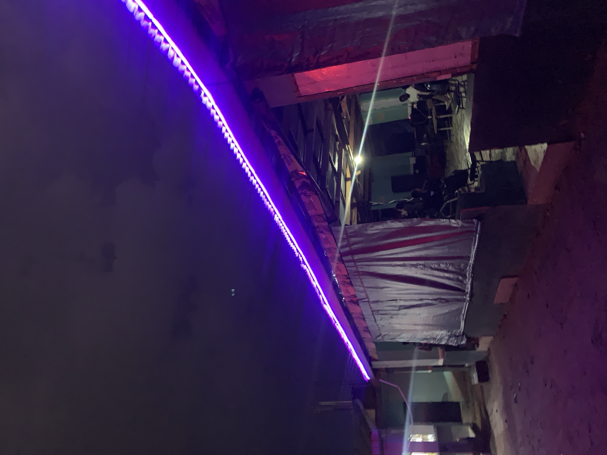 An outdoor gathering is taking place at night in Jos, Nigeria, under a cloudy sky. A building with a corrugated metal roof is illuminated by a string of vibrant purple LED lights running along the overhang. The building appears to be an open-air establishment, with a large, dark tarp or curtain hanging down on the left side. Inside, several individuals are visible, some seated at tables and others standing. The ground in front of the establishment is a dirt or gravel surface, partially covered in what appears to be more purple lighting or reflection from the LEDs. To the left of the building, there is another structure with a metal roof, and a vertical pole. The overall atmosphere suggests a casual, possibly festive, nighttime event.