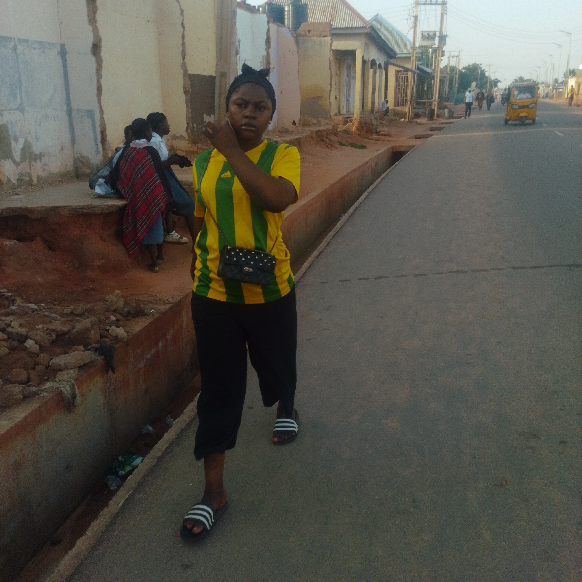 In an urban environment, a young woman with dark skin and black hair, wearing a black bow, walks on a paved street. She is dressed in a yellow t-shirt with green stripes and black pants. A black purse with white studs is slung across her body. Her feet are adorned with black and white striped slides. Behind her, a group of children is visible, with one child in the foreground wearing a red and white plaid shawl. The backdrop includes dilapidated buildings and a drainage ditch containing debris. In the distance, a yellow auto-rickshaw and several other pedestrians are present on the street. The scene appears to be an outdoor street view.
