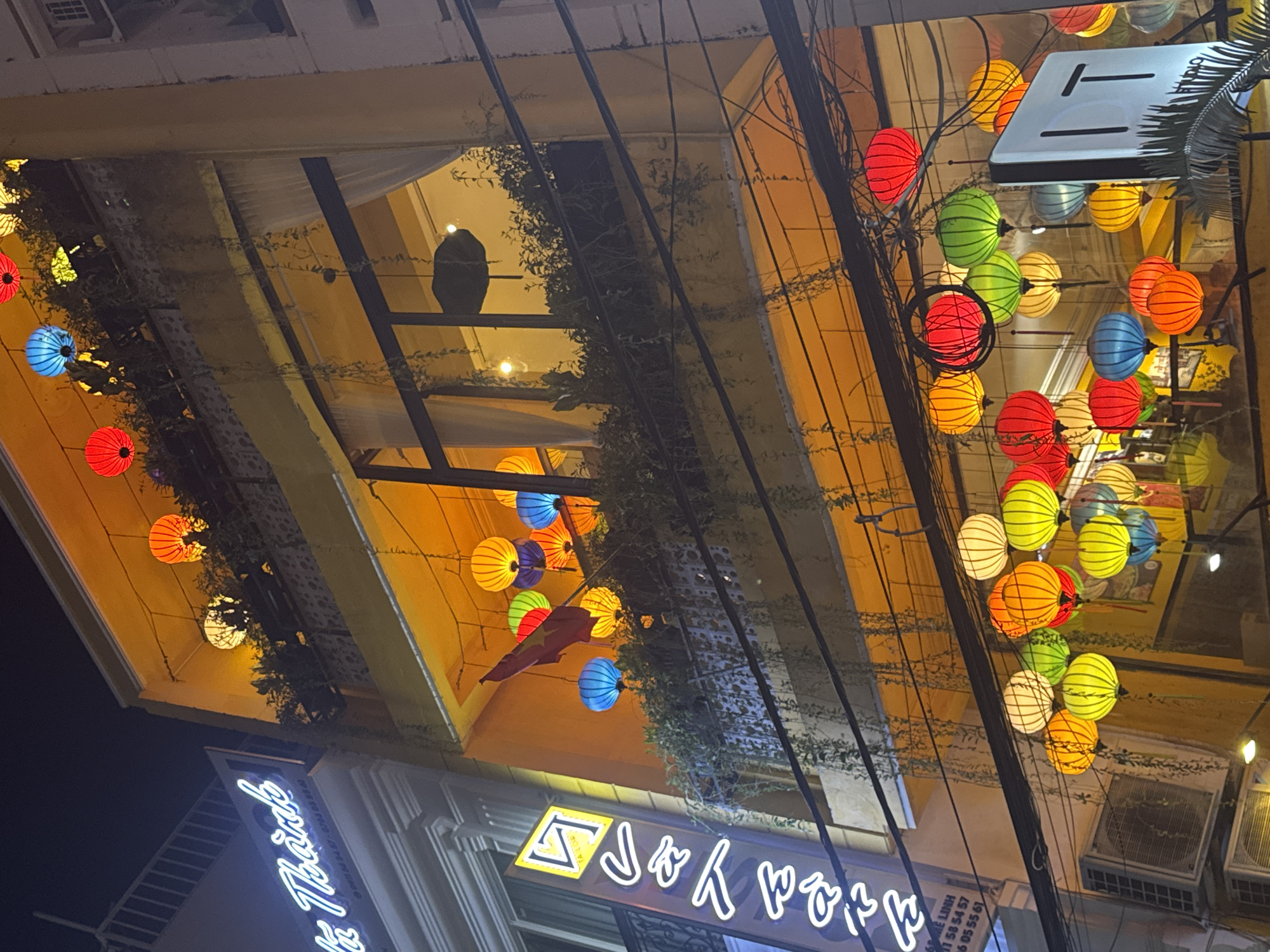 Captured at night in Nha Trang Ward, Vietnam, the image shows buildings adorned with strings of colorful lanterns. The lanterns are predominantly red, green, yellow, and blue, casting a warm glow. Dense clusters of foliage, likely vines, are visible against the yellow awnings of the buildings. Overhead power lines crisscross the view. Below the awnings, signage with neon lighting spells out "Sky Park" and "The Tide." The architecture suggests a commercial area with bars or restaurants.