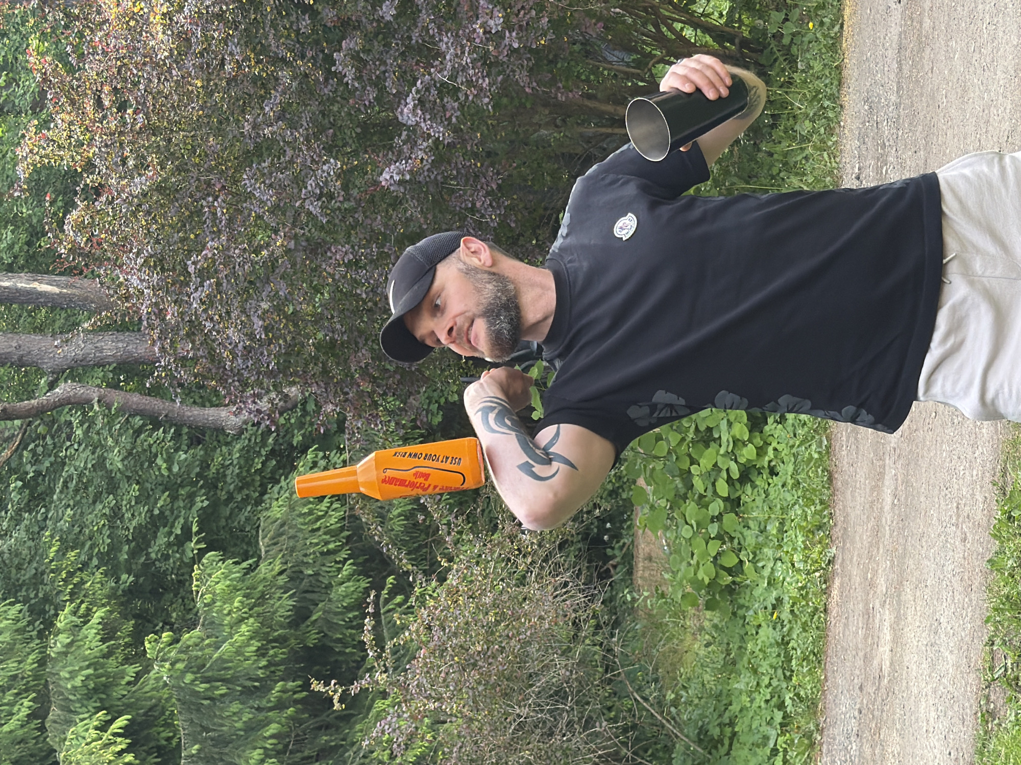 A bearded man wearing a black baseball cap, a black t-shirt with a grey floral pattern, and light-colored shorts stands outdoors, balancing an orange bottle on his right forearm. He holds a metallic cup in his right hand. The background consists of lush green foliage, trees, and a dirt path. The scene is an outdoor setting, possibly a park or a backyard, with abundant vegetation. The lighting suggests it is daytime, with sunlight filtering through the leaves. There is no indication of the city of Tallinn, Estonia, within the image.