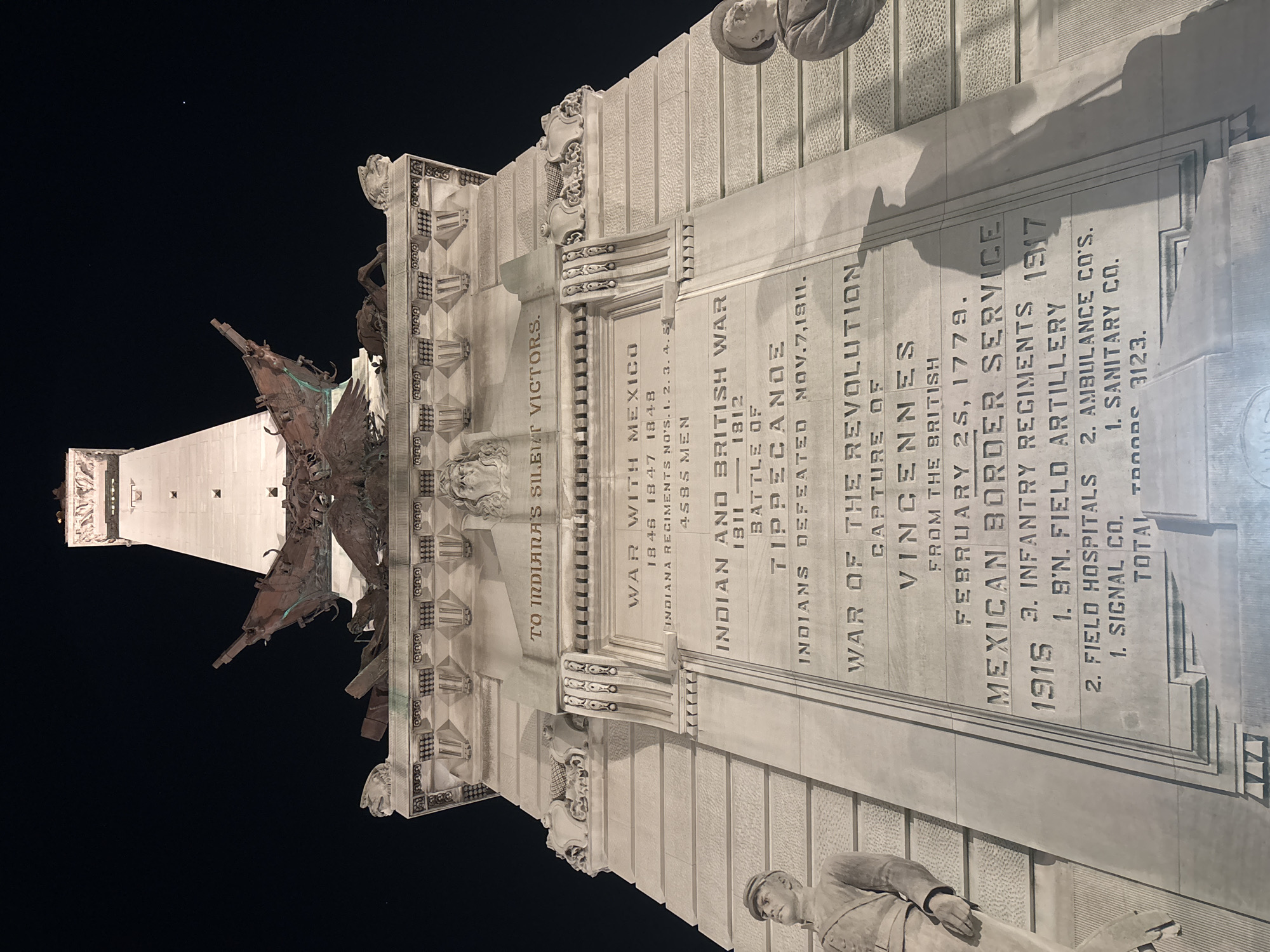 The image depicts the Indiana World War Memorial in Indianapolis, United States of America. A towering white monument dominates the night sky. In the foreground, a portion of the memorial's base is visible. It features stone carvings, including a plaque with the inscription "TO INDIANA'S SILENT VICTORS" and listing various wars and military engagements: the War with Mexico, Indian and British War (Battle of Tippecanoe), War of the Revolution (Capture of Vincennes), and Mexican Border Service. These inscriptions indicate the wars in which soldiers from Indiana served. A carved soldier is also visible.