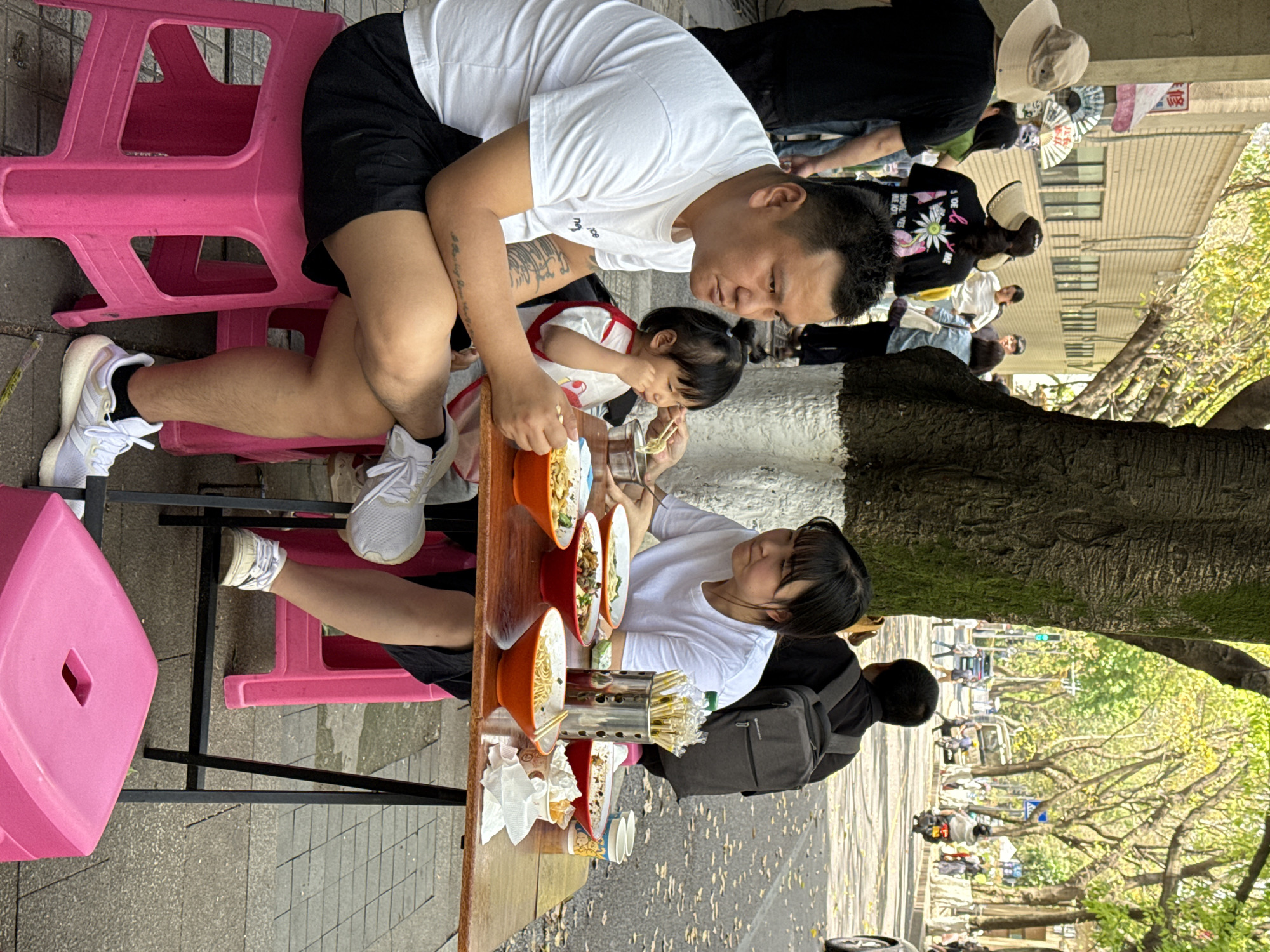 In Yuzhong District, China, a family is eating at a small outdoor noodle stand. A man in a white t-shirt and black shorts sits on a pink plastic stool, helping a young child with their noodles. A woman in a white t-shirt sits on another pink stool across from them. Several bowls of noodles are on the table. A large tree is visible in the background, and other people are walking by on the sidewalk.
