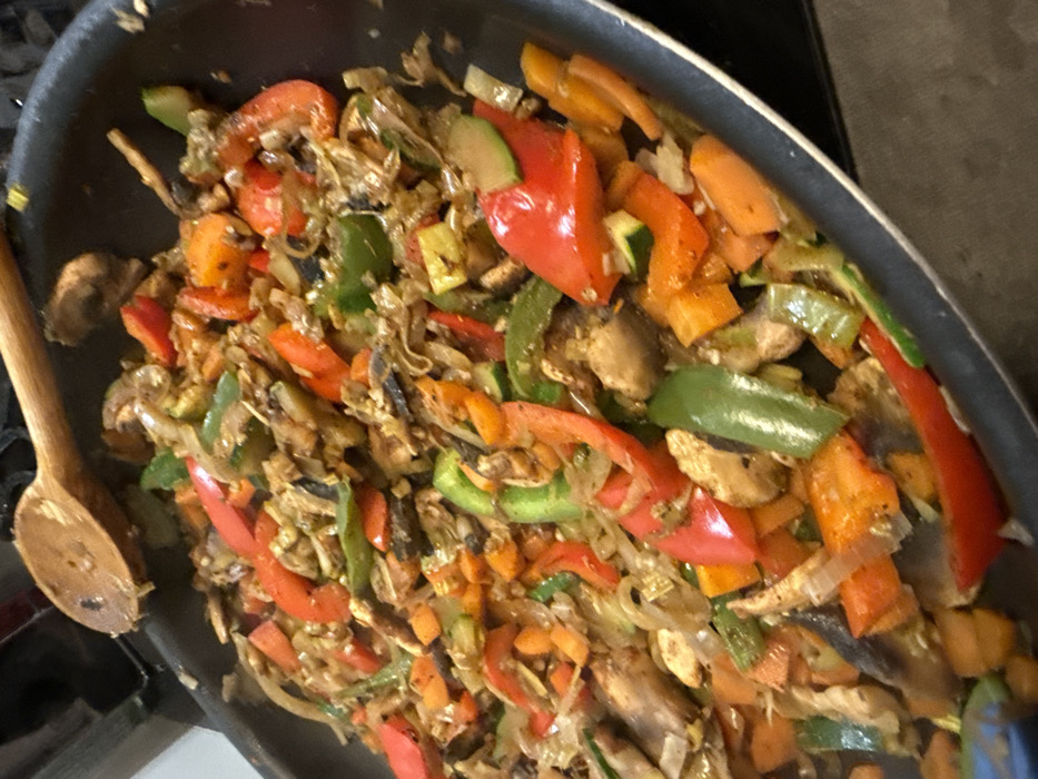 The scene depicts a close-up, elevated view of a large black frying pan or wok filled with a stir-fried vegetable mixture, located within a kitchen setting in Strasbourg, France. The pan contains a vibrant assortment of chopped ingredients, including prominent pieces of red, green, and orange bell peppers, sliced mushrooms, translucent onion slices, and rounds of zucchini. Smaller, diced orange pieces, likely carrots, are also interspersed throughout. The vegetables appear cooked, with some exhibiting a slight charring or caramelization, and are coated in a visible sheen, suggesting oil or sauce. A light-colored wooden spoon with a rounded head rests partially submerged in the upper left section of the pan, indicating recent or ongoing stirring. No individuals are visible. The background is blurred and dark, consistent with a stovetop and immediate kitchen environment.