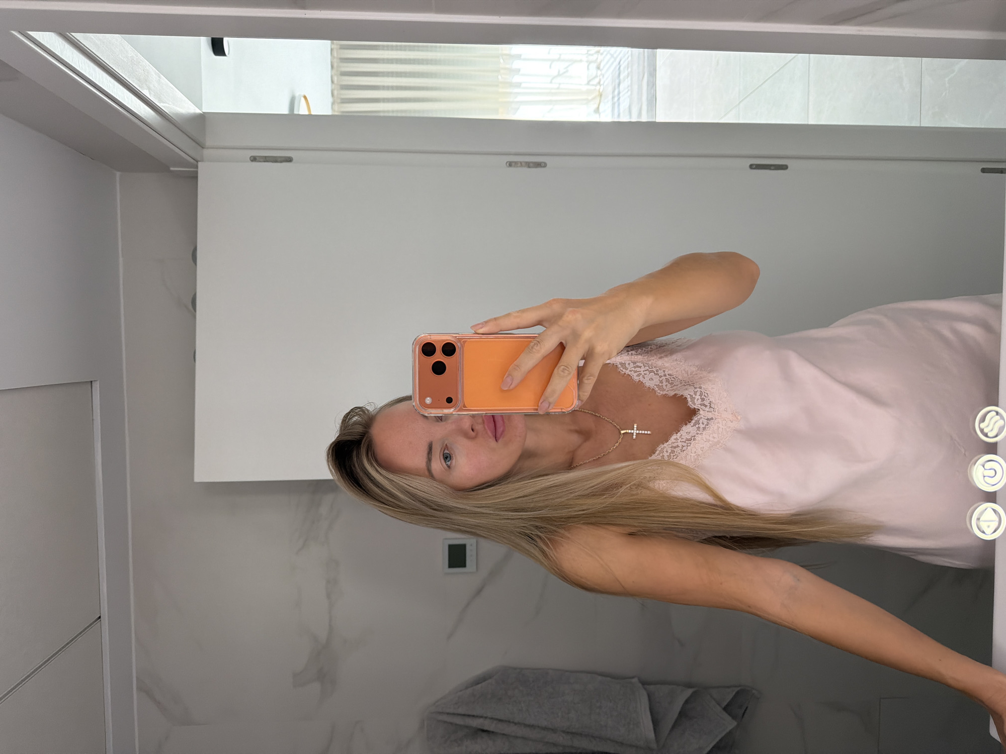 A woman with long blonde hair takes a selfie in a bathroom. She is wearing a light pink slip with lace trim and a silver cross necklace. Her right hand is holding an orange-cased phone, and her left arm is resting on a white surface. The bathroom features marble-patterned walls, a grey towel hanging on the left, and a door on the right that opens to a room with sheer curtains and tiled floors. The scene is set indoors, likely in a residential or hotel bathroom. The prompt indicates the location is in Finestrat, Spain, but this cannot be visually confirmed from the image alone.