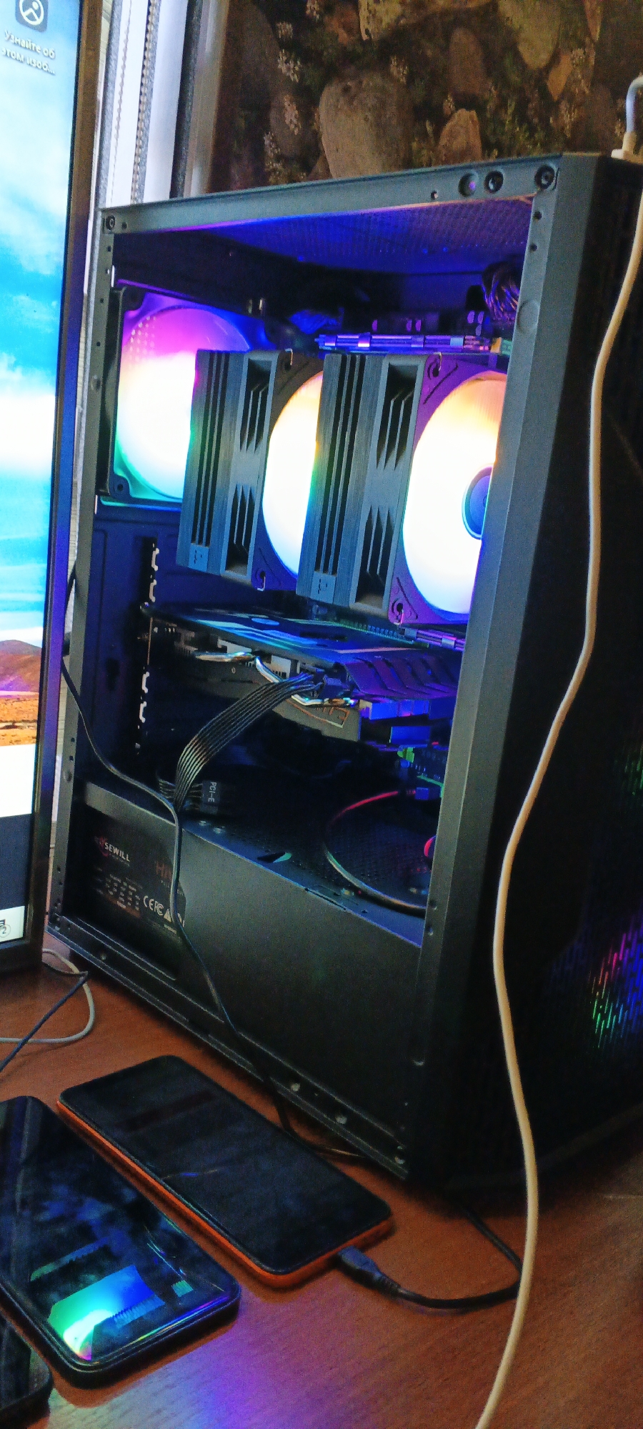 The image displays the interior of a computer case, illuminated by RGB lighting. Prominently featured are three large computer fans with colorful lighting, two of which are partially obscured by heatsinks. Below the fans, a graphics card is visible, connected by black cables. A power supply unit with "SEWILL" and "CEFC A" branding is situated at the bottom of the case. The computer case itself is black. In the foreground, two smartphones are visible on a wooden surface, with one of them plugged in and charging. The background shows a wall with a floral wallpaper and a portion of a computer monitor displaying an image of a landscape. The overall scene is indoors, with artificial lighting creating a colorful ambiance. There is no indication of the location being Mustakillik, Uzbekistan.