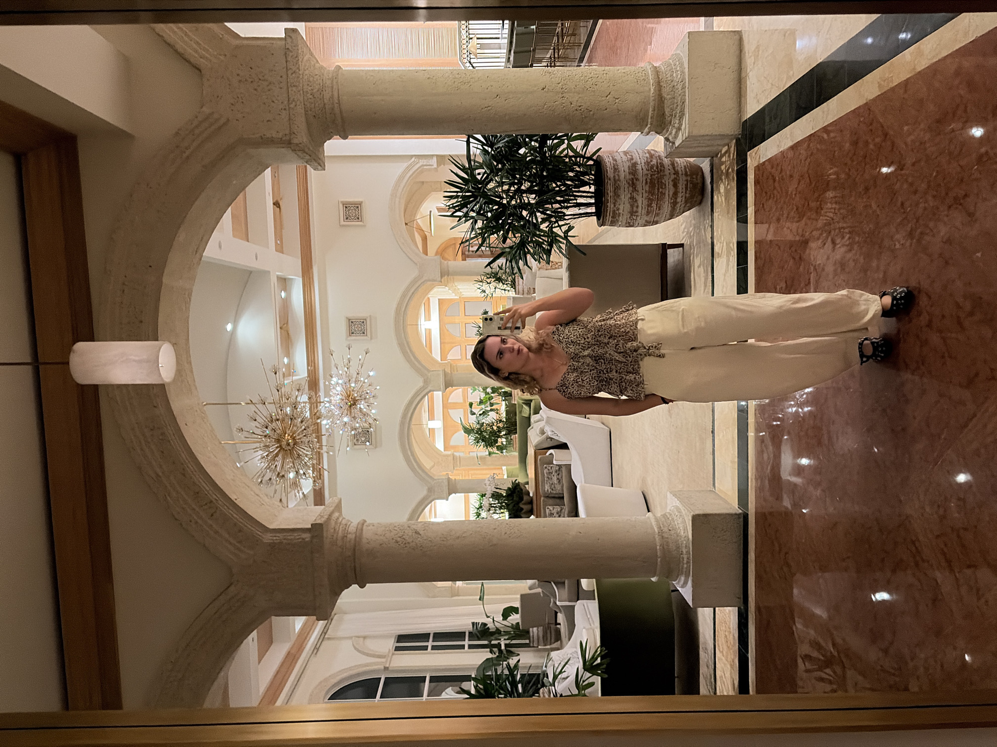 The photo depicts a woman taking a selfie inside a grand building, possibly a hotel or resort lobby. The architectural style features ornate columns, arches, and decorative chandeliers. Potted plants are placed throughout the space. The floor is made of polished marble in various colors, creating a patterned effect. Furniture is seen in the background, including sofas and chairs. The location is identified as Key Biscayne, United States of America.