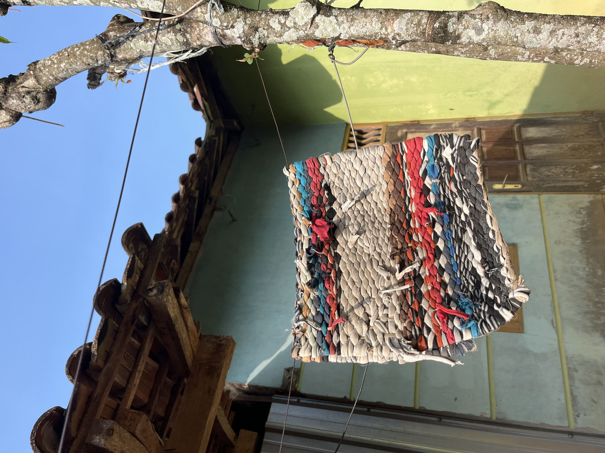 A handmade rug, composed of various colored fabric strips woven together, hangs from a clothesline suspended between a tree and a building. The rug displays a colorful pattern with dominant hues of red, blue, black, white, and beige. It appears worn and somewhat frayed. The background consists of a light green building with a brown wooden door and a tiled roof. To the right, the textured bark of a tree is visible, with some twine wrapped around it. The scene is illuminated by bright sunlight, casting shadows. The location is identified as Glagah Kulon, Indonesia, suggesting a rural or suburban setting. There are no people or animals present in the image.