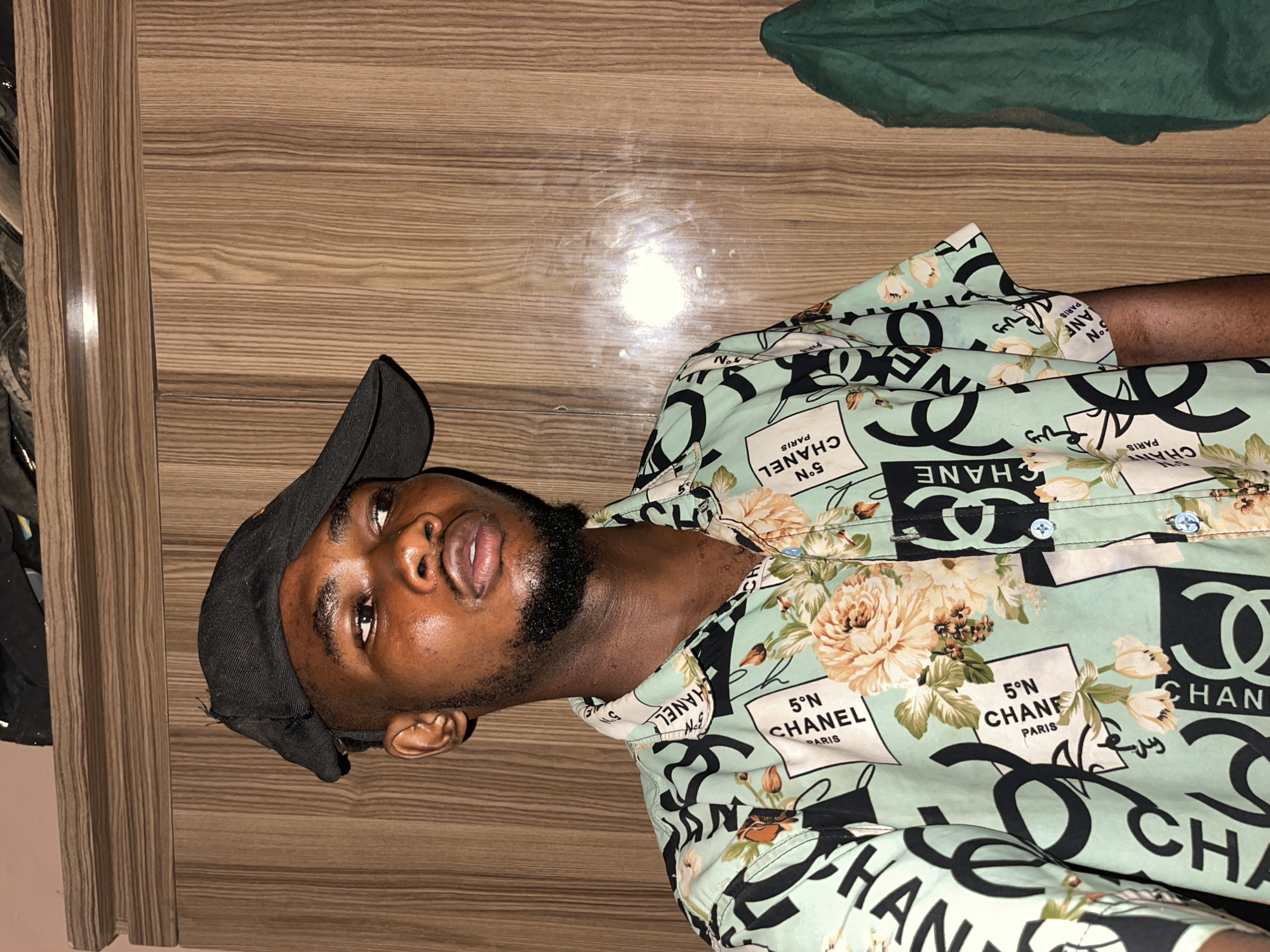 A medium shot captures a Black man, oriented vertically, against a backdrop of wood paneling and a green object in the upper right corner. The man wears a black baseball cap, tilting slightly to his left. He sports a black beard, and his skin appears shiny. He is attired in a button-down shirt with a light green base, decorated with Chanel logos and floral designs. The wood paneling features horizontal lines and a glossy finish. The lighting appears to be artificial. No location data is discernible from the image. Unguwar Gabas, Nigeria cannot be determined from the visual information in the image.