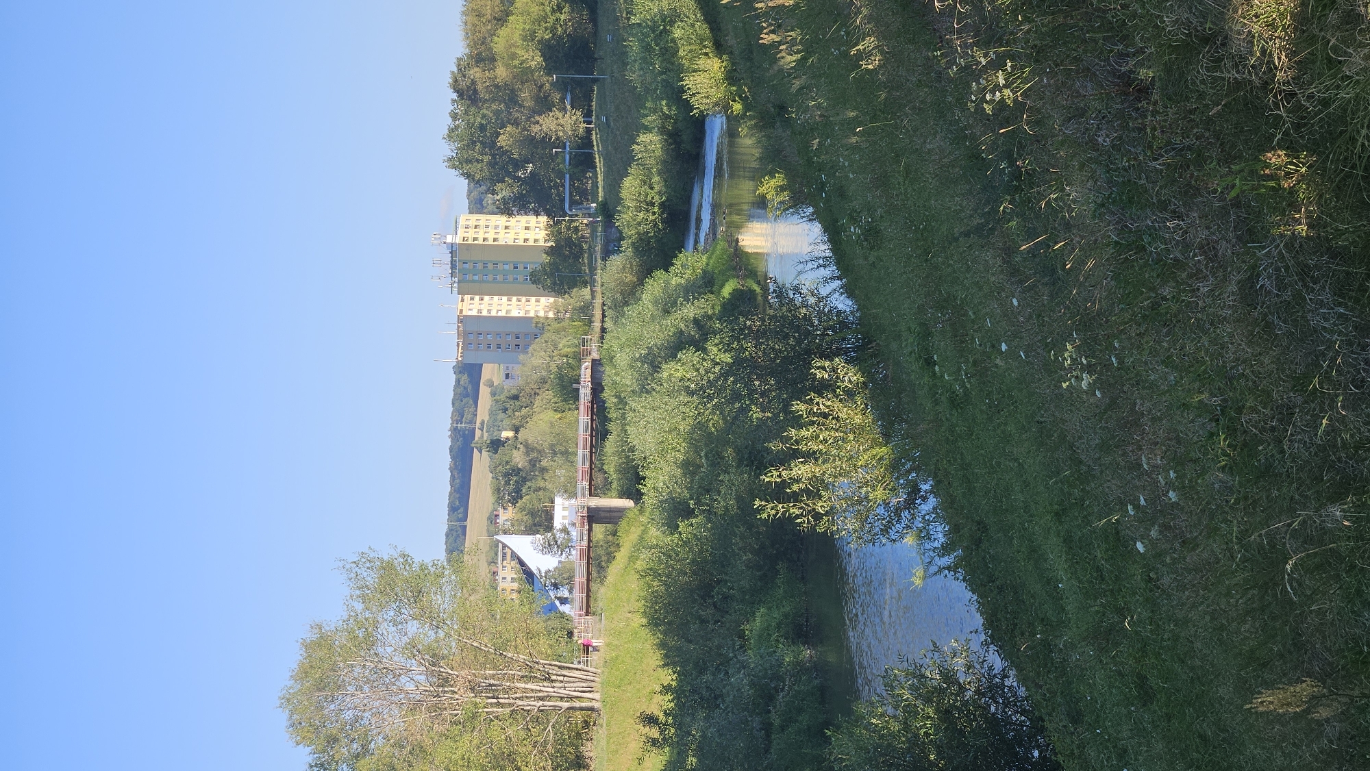 The image captures a sunny daytime scene in Prešov, Slovakia, likely depicting the area near the Sekčov river. In the background, a tall, light green apartment building stands prominently, characterized by its numerous windows and a telecommunications tower on its roof. To the left of this building, a smaller, white and blue structure with a distinctive angled roof is visible. A bridge, appearing to be made of metal with concrete supports, spans the river in the midground. One person is partially visible walking on the bridge, dressed in pink. The river itself flows gently through the scene, reflecting the blue sky and surrounding greenery. The foreground and riverbanks are covered in lush, green vegetation, including trees, bushes, and tall grasses, suggesting a natural or park-like setting within the city. The sky is a clear, bright blue, indicating fair weather.
