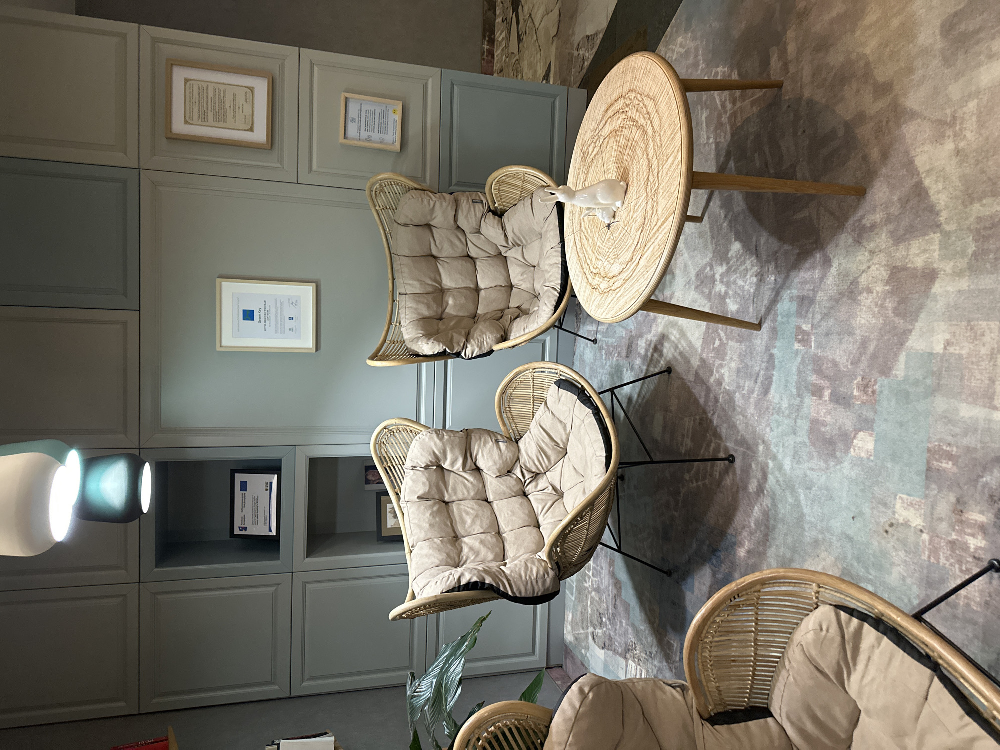 The image captures an interior space with notable design elements. Dominating the scene are three rattan chairs, each furnished with tufted cushions. Two of the chairs are positioned closely together, while the third is partially visible in the lower right corner of the image. A round wooden table stands beside one of the chairs, hosting a decorative white bird figure.
The chairs and table are set against a backdrop of a painted wall and a built-in shelving unit. The wall is adorned with framed certificates or decorative papers. The flooring displays a mottled pattern in muted tones of gray and brown.
The scene is bathed in natural light, contributing to the space's bright and airy ambiance. The overall impression is one of a well-designed, comfortable, and inviting interior space. The location is Wrocław, Poland.