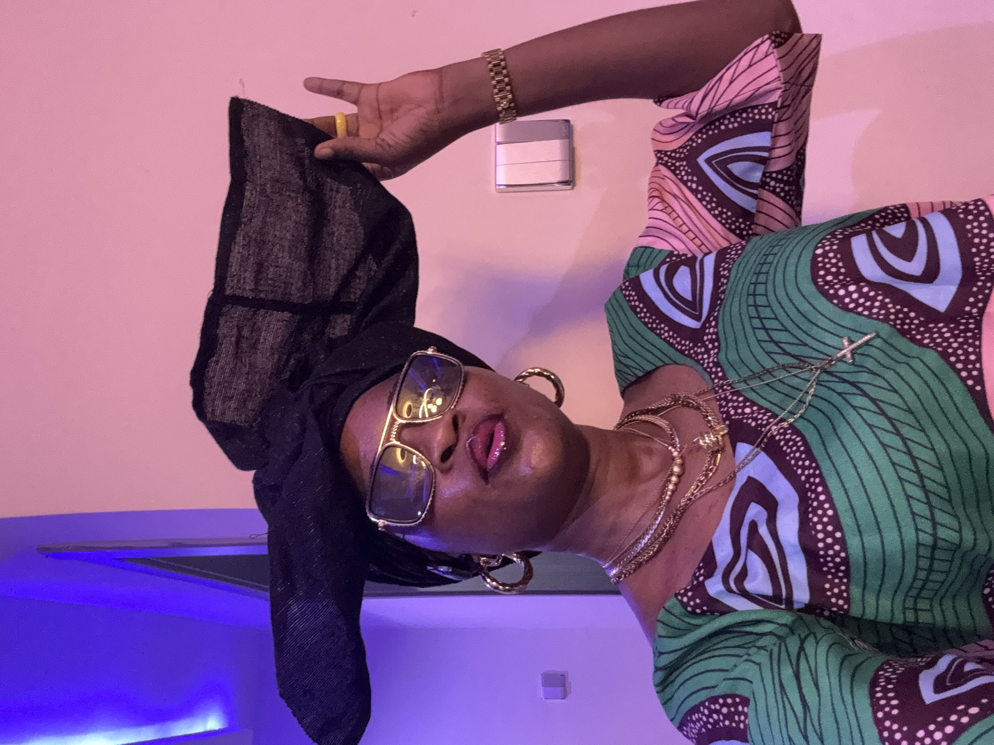 The image shows a woman wearing a black headwrap, large gold-rimmed glasses, and a multi-colored patterned dress. She is also adorned with multiple gold necklaces, large hoop earrings, a yellow ring, and a gold wristwatch. The woman is holding the edge of her headwrap with her right hand. The background is a pink wall, with a light fixture casting a purple and blue light on the left side of the image. There is a light switch on the wall behind her right arm. The image appears to have been taken indoors in Ajara, Nigeria.