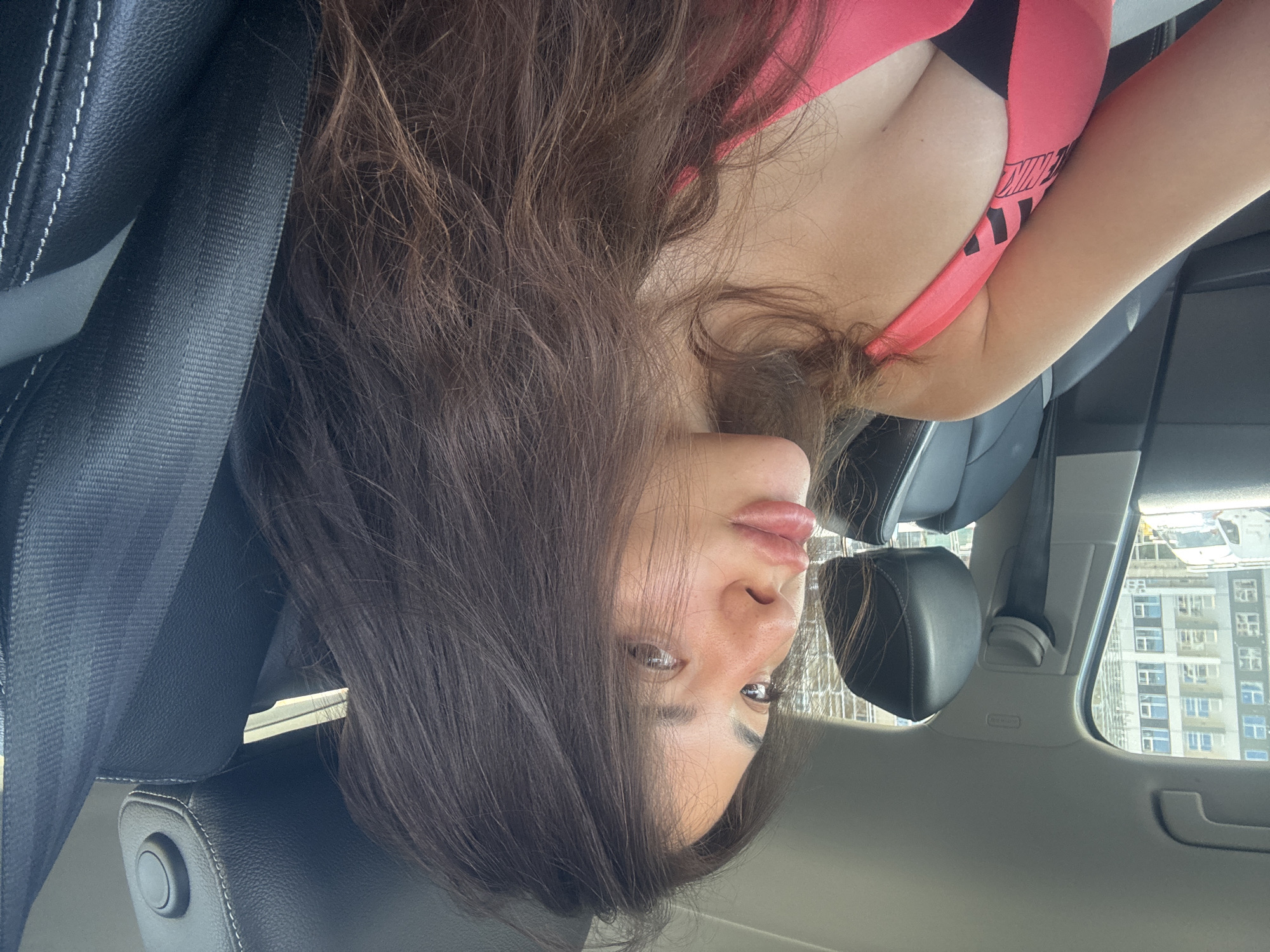 A young woman with long, dark brown hair is seated in the passenger seat of a car, looking to her left. She is wearing a pink sports bra with black accents and what appears to be a black undershirt. Her hair is partially obscuring her face and shoulders. The interior of the car is visible, including a black leather seat with white stitching, a seatbelt, and the headrest. Outside the car window, a multi-story building with numerous windows is visible, suggesting an urban environment. The scene is likely in Plovdiv, Bulgaria, given the architectural style of the building which resembles those found in modern developments in the city. The lighting suggests it is daytime.