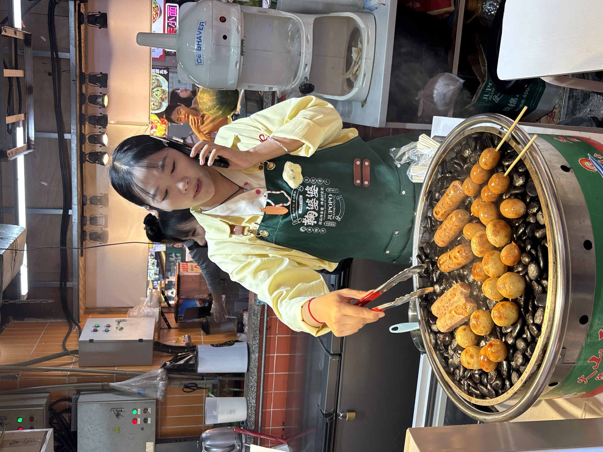 A medium, eye-level, indoor shot shows a young female street vendor in Yuzhong District, Chongqing, China, operating a food stall. She is facing left with her head turned towards the camera, talking on a smartphone and using red tongs to turn food on a round grill. The grill has round, golden food items and sausages. The vendor is wearing a light yellow shirt and a green apron that has "JUPO" in white letters. Above her on the left is a row of overhead lights. Behind the grill, there is an ice shaver machine, other restaurant customers, and a menu board. The background includes a red tiled wall and various appliances and food containers.