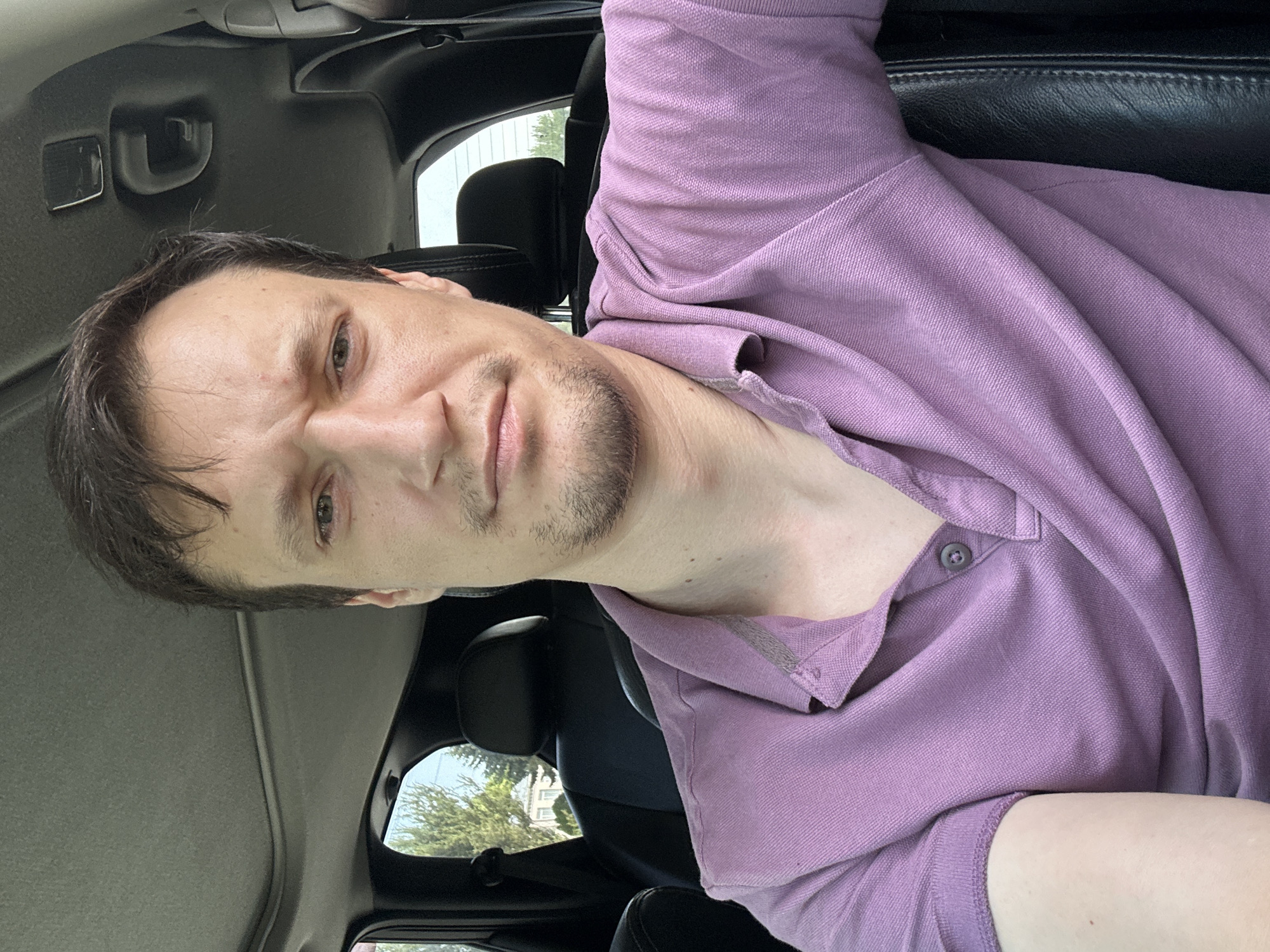 A medium shot selfie shows a man sitting in the driver's seat of a vehicle. The man has fair skin, brown hair, and light-colored eyes. He is wearing a purple collared shirt with a button closure. His expression is neutral, with his eyebrows slightly furrowed. The background shows the interior of the car, including black leather seats and a sunroof. Through the windows, trees and a building are visible, suggesting an outdoor setting within a city or suburban area. The image is taken during daylight hours. The location is not specified in Makiv, Ukraine.