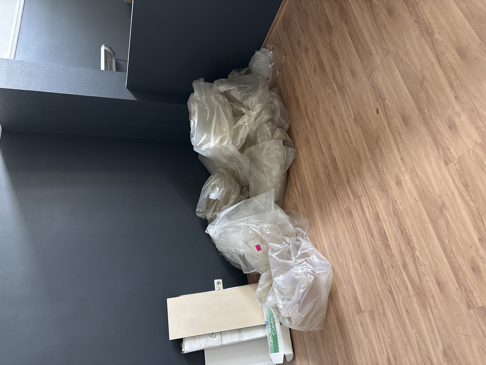 In Zaporizhzhia, Ukraine, a room with dark gray walls and a light wood-grain linoleum floor contains a pile of plastic-wrapped items and some flat boards leaning against the wall. The plastic-wrapped items are in large, translucent bags, appearing to be upholstery or padding. A pink sticker is visible on one of the plastic bags. Among the stacked items, there are at least three flat wooden boards, one of which has a white sticker with a black number six. A white electrical outlet is visible on the wall behind the boards. The lighting suggests it is daytime, with light casting shadows on the wall from an unseen source. The scene appears to be an interior space that is either under renovation or being prepared for storage.