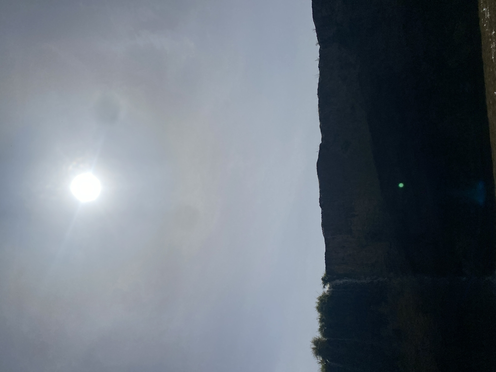 The image displays a dramatic natural landscape in Maigizo, Nigeria, featuring a large cliff or mountain under an overcast yet bright sky. The sun, a brilliant white sphere, is prominently located in the upper left portion of the frame, emitting strong light and causing a pronounced lens flare. The sky is a light grey, indicating significant cloud cover, but the sun's intensity confirms it is daytime.
A massive, dark geological formation, appearing to be a cliff or mountain face, dominates the right side of the image, extending vertically from the bottom to the top. Its surface is rugged and predominantly dark, suggesting it is either in shadow or composed of dark rock. Sparse vegetation, possibly small trees or bushes, clings to the edges and base of this imposing structure. A distinct green, circular lens flare is visible within the darker expanse of the cliff, an optical artifact from the direct sunlight.
There is no discernible activity or event taking place, and no people or visible text are present. The image focuses entirely on the contrast between the radiant, overcast sky and the stark, dark landform, emphasizing the raw power of nature in this Nigerian setting.