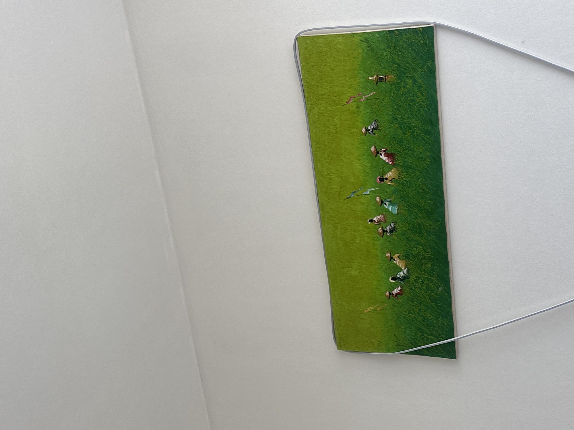 A vertically oriented photograph depicts a wall corner in North Kuta, Indonesia, where a rectangular, framed picture hangs. The artwork features a green field populated with small figures, possibly depicting farmers working the land. The frame is light gray, and a thin white wire is attached, presumably for hanging. The walls are white with a slight texture.